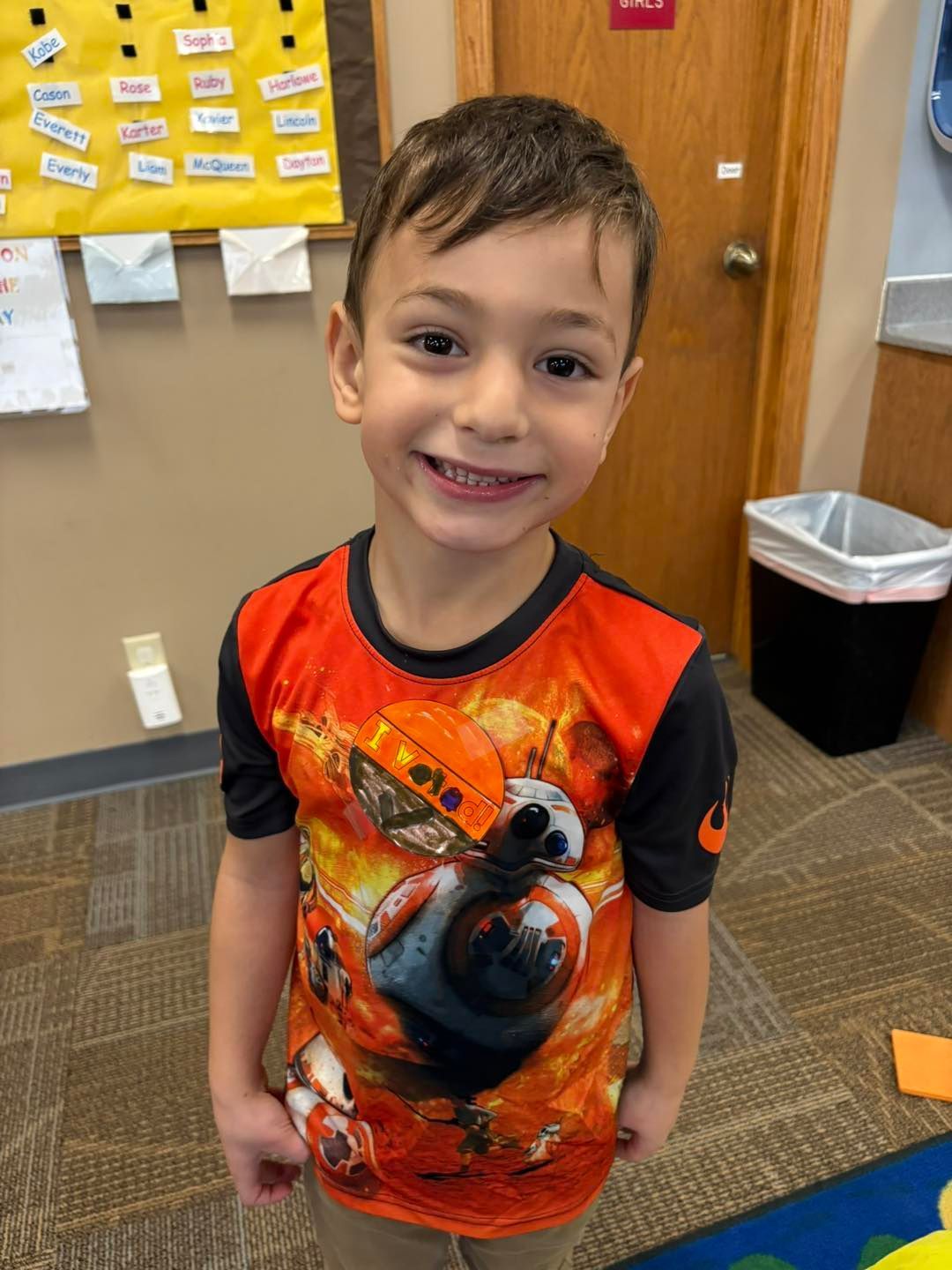 A young boy wearing a star wars shirt is smiling in a classroom.