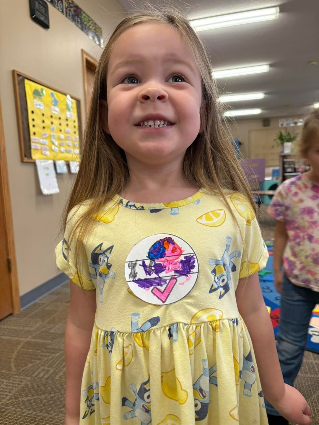 A little girl in a yellow dress is smiling in a classroom.
