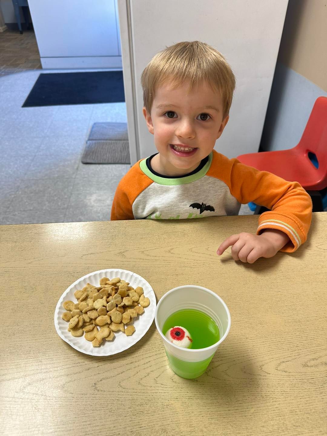 A young boy is sitting at a table with a plate of crackers and a cup of green liquid.