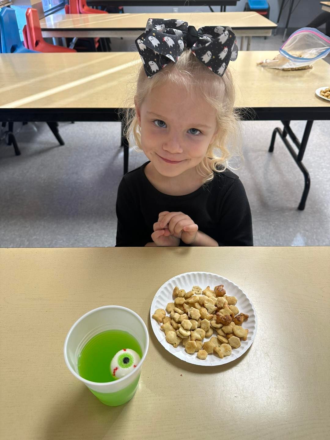 A little girl is sitting at a table with a cup of green liquid and a plate of snacks.