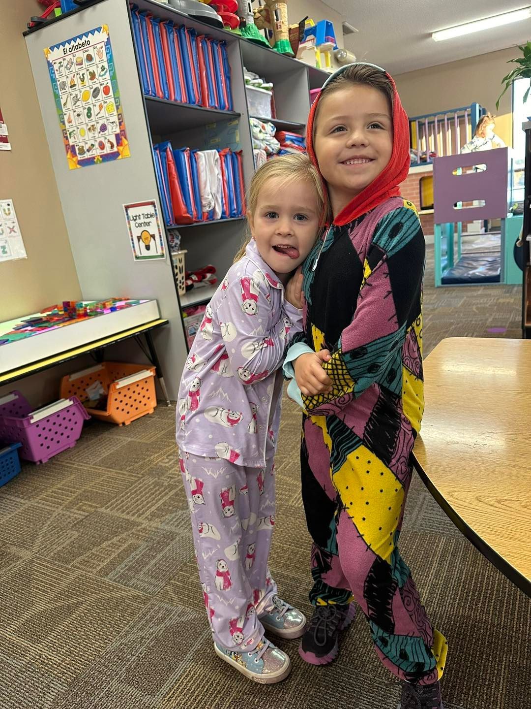 Two little girls in pajamas are hugging each other in a classroom.