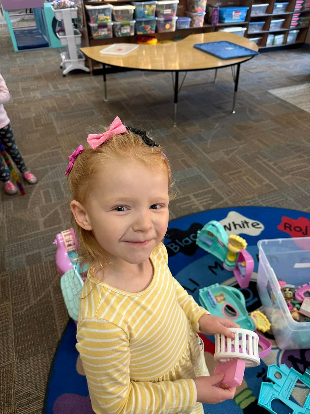 A little girl in a yellow dress is smiling in a classroom.