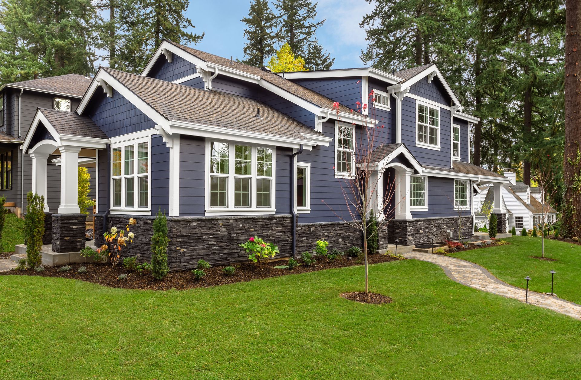 Blue house with white trim, stone base, and green lawn. Pathway leads to the front door; trees in the background.