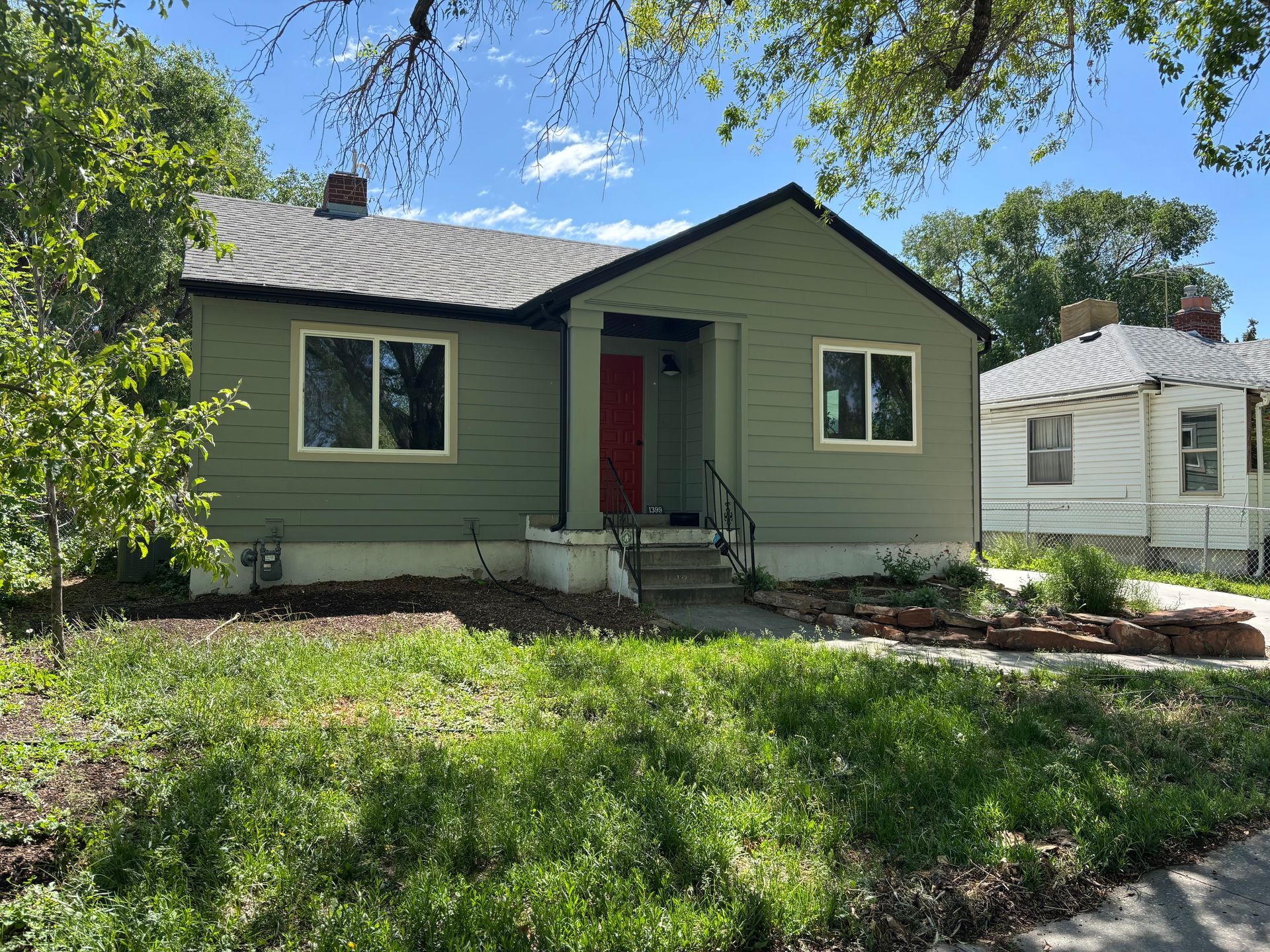 Green house with red door and light trim, lawn in foreground, neighboring house to the right.