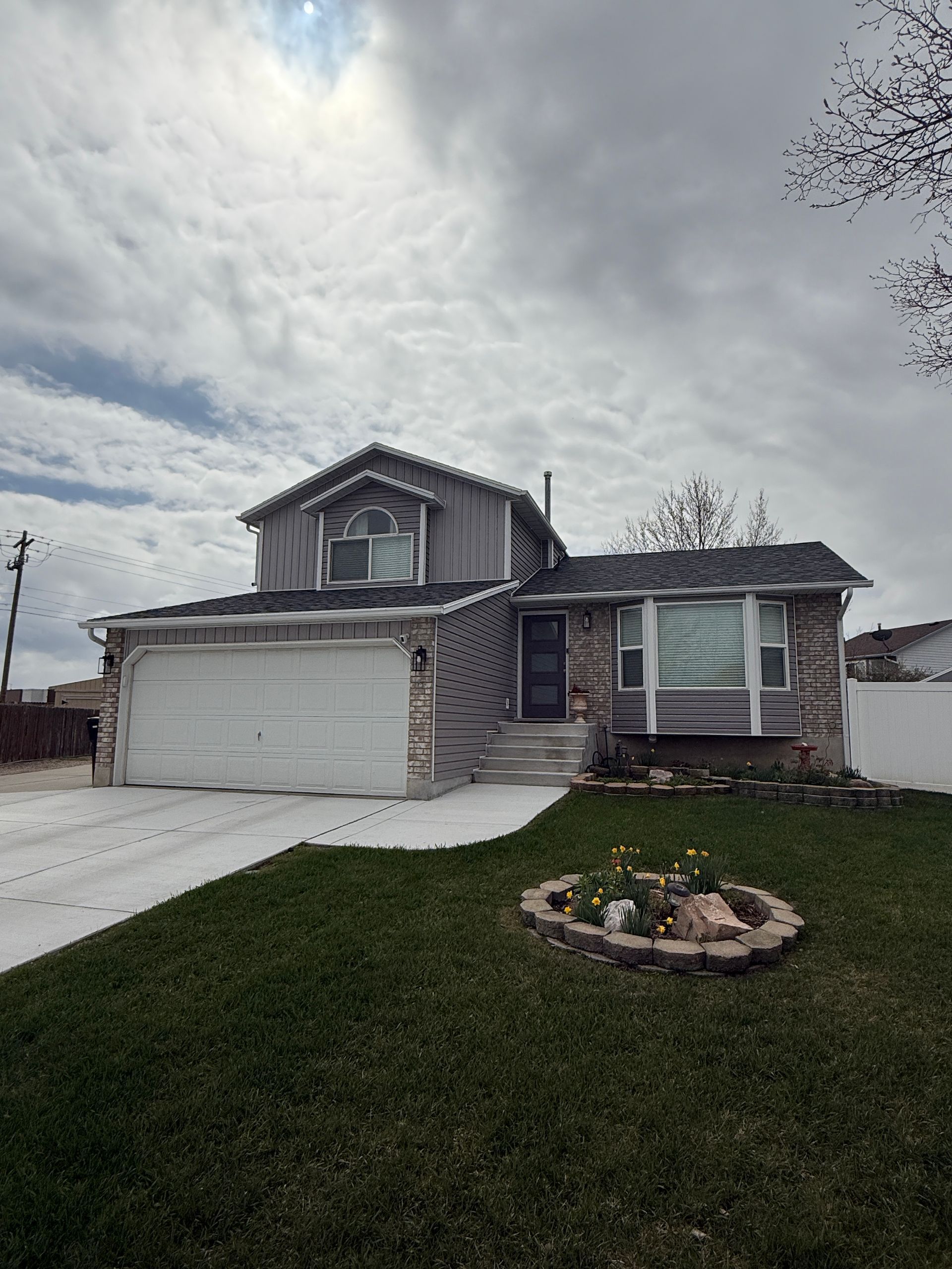 Two-story house with gray siding and a white garage door, on a grassy lawn under a cloudy sky.