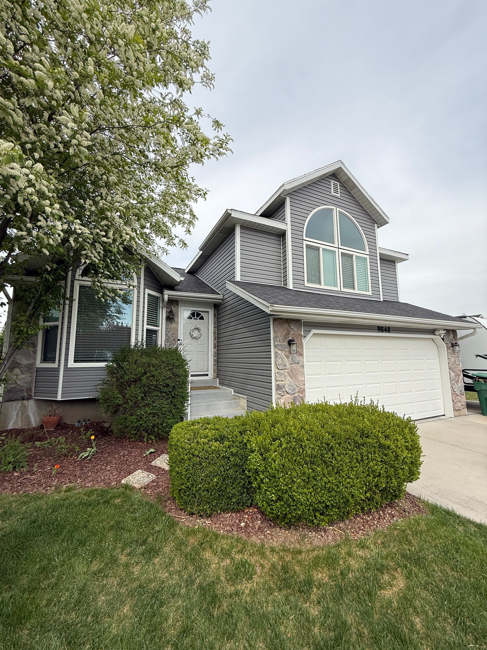 Gray house with white trim, arched window, and attached garage. Green lawn, shrubs, and tree.