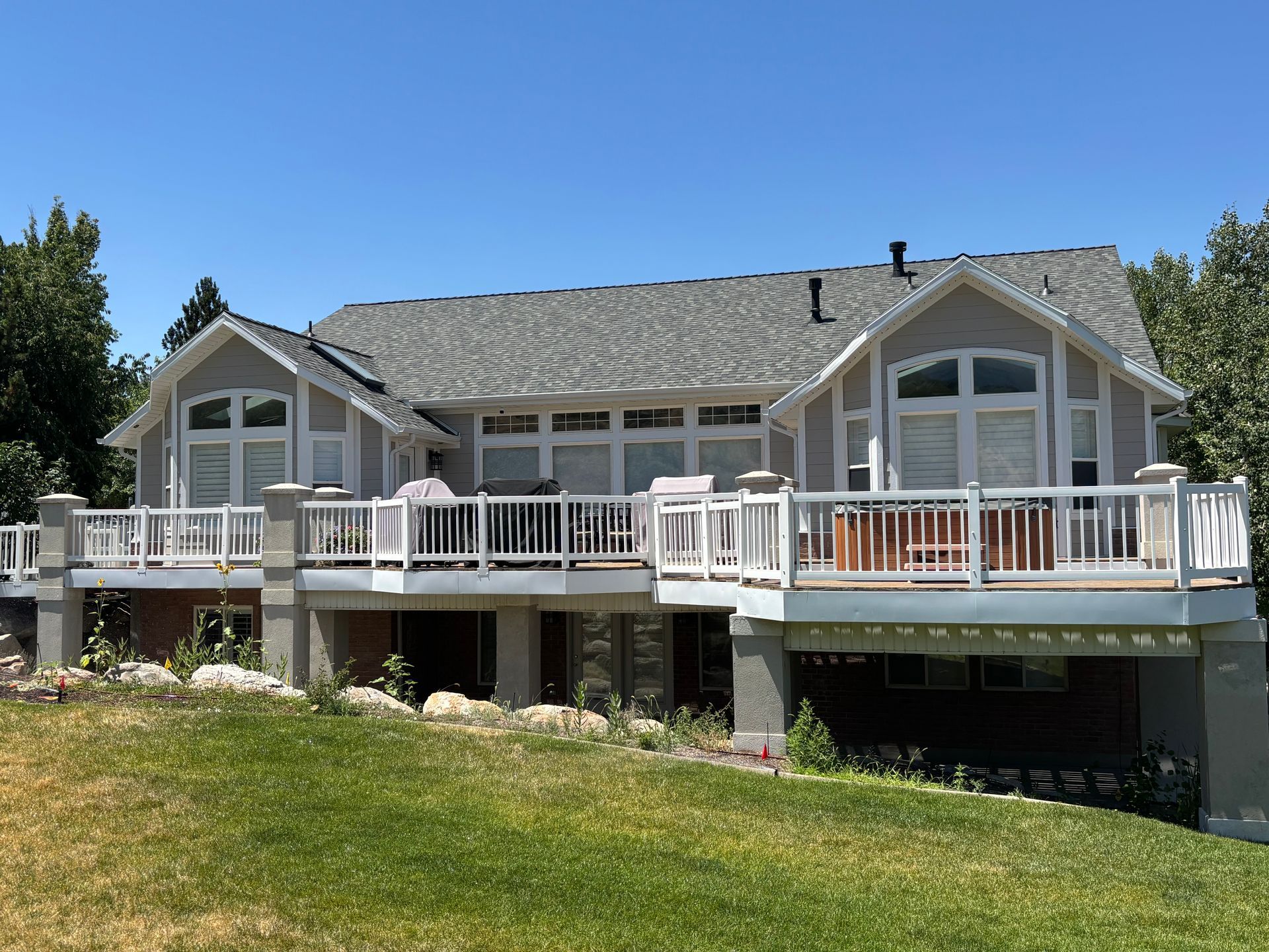 Two-story gray house with large windows, white railings, and a deck, viewed from a grassy yard on a sunny day.