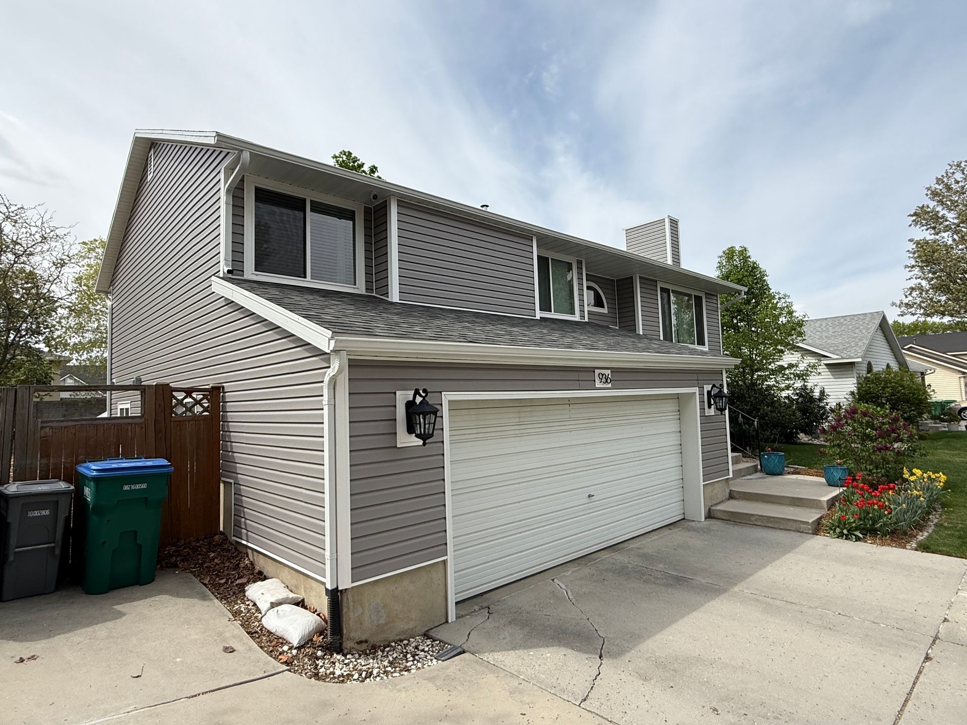 Two-story gray house with white garage door and trim, blue recycle and black trash cans, cloudy sky.
