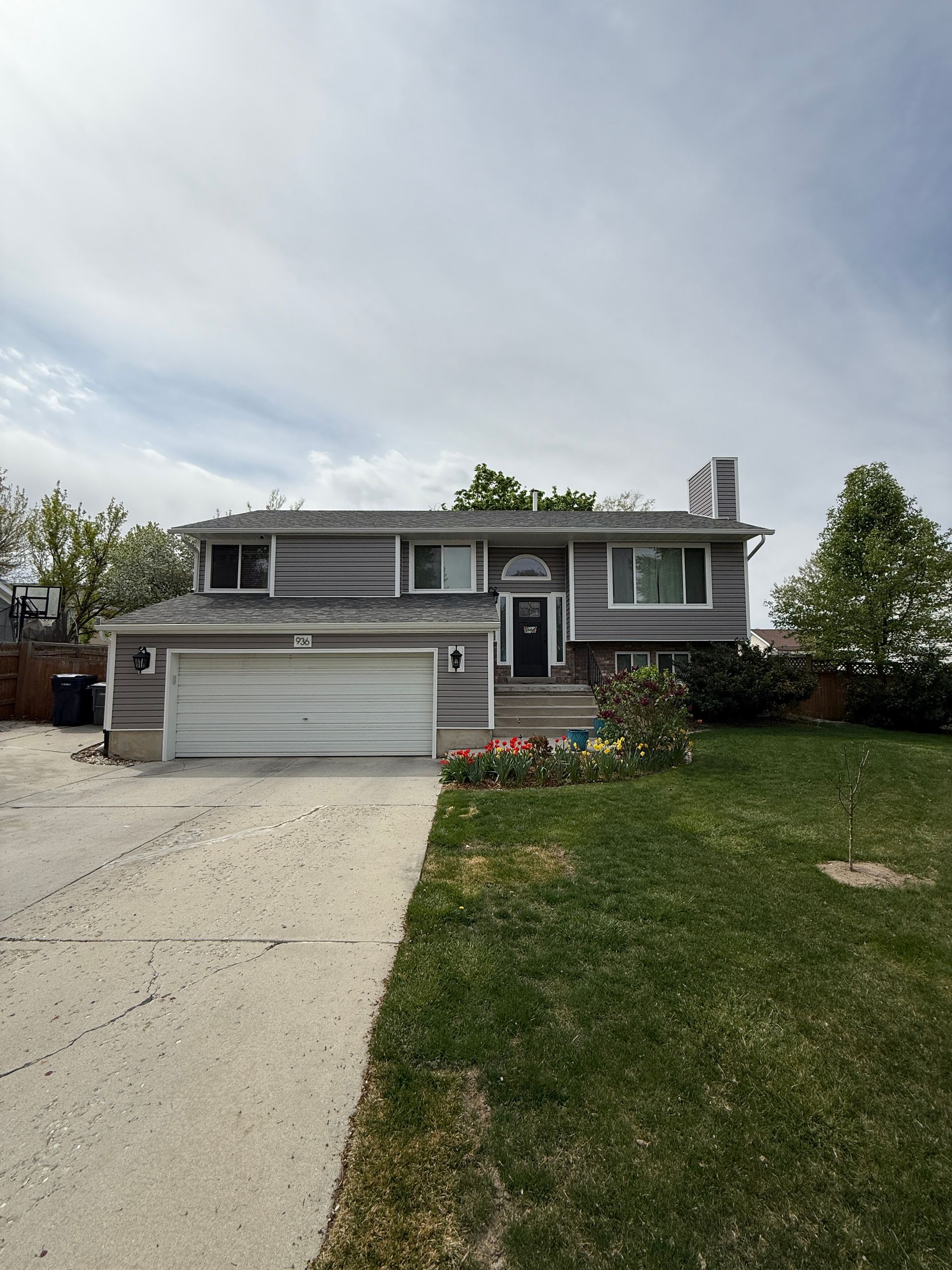 Gray two-story house with a driveway and green lawn under a cloudy sky.