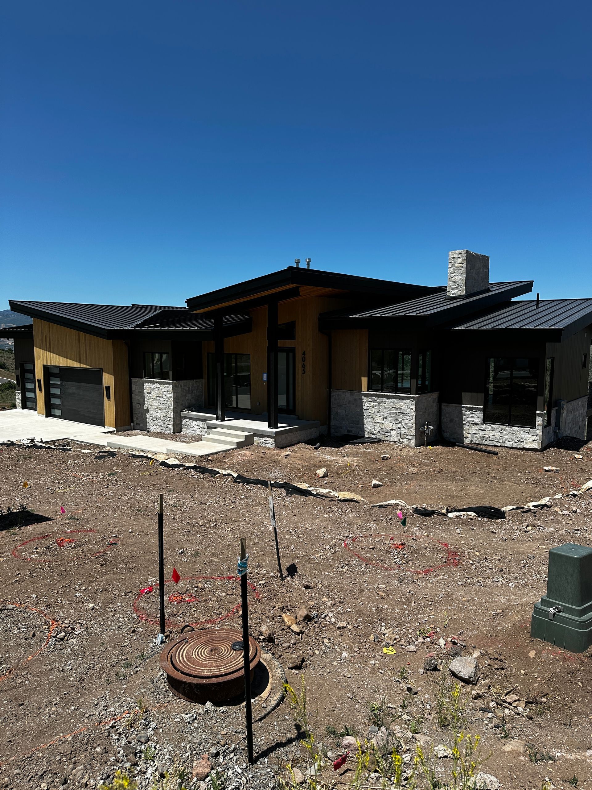 Modern home with stone accents, black roof, and a dirt yard under a blue sky.