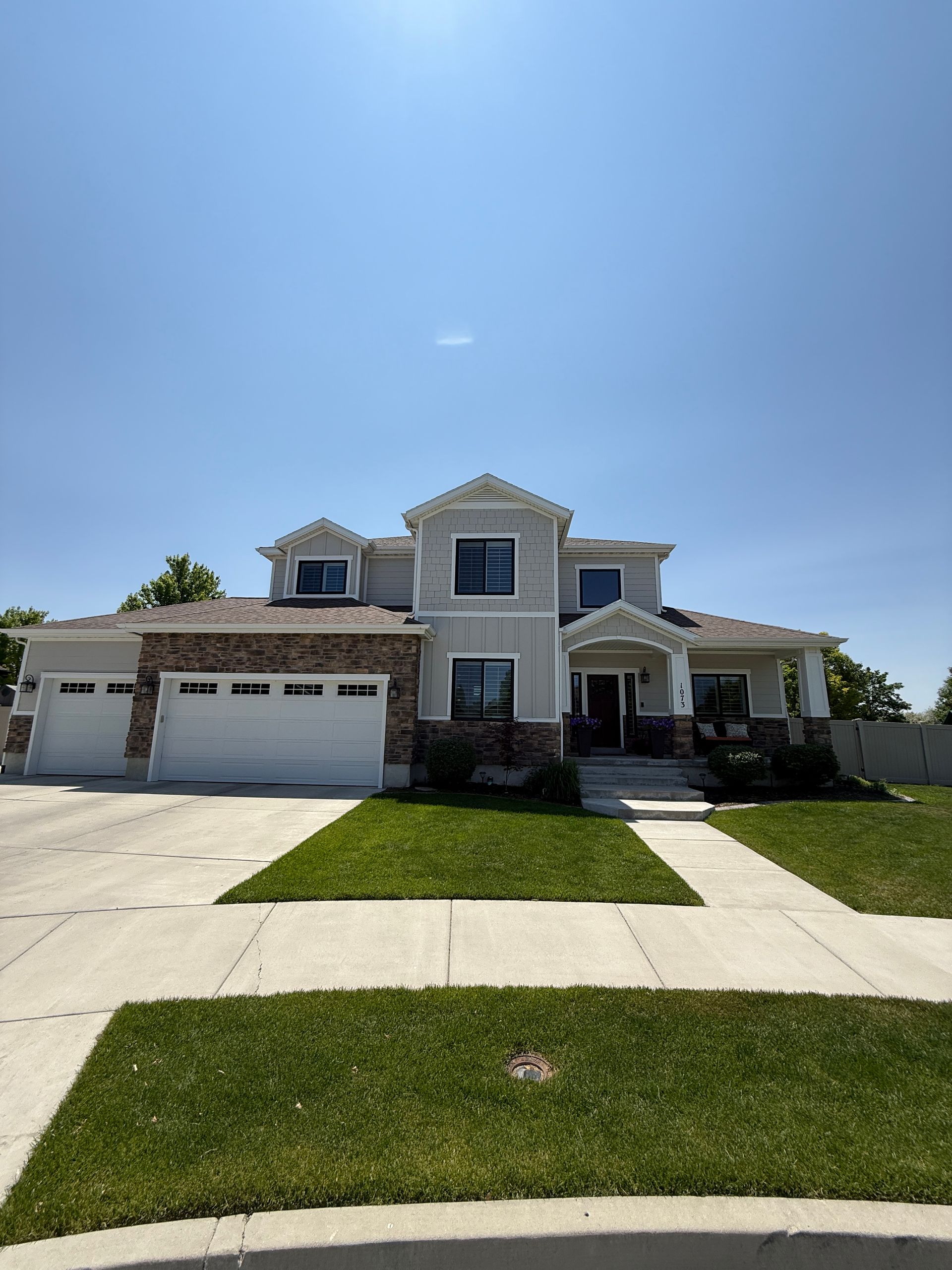 Two-story house with grey siding, stone accents, white garage doors, and a green lawn under a sunny sky.