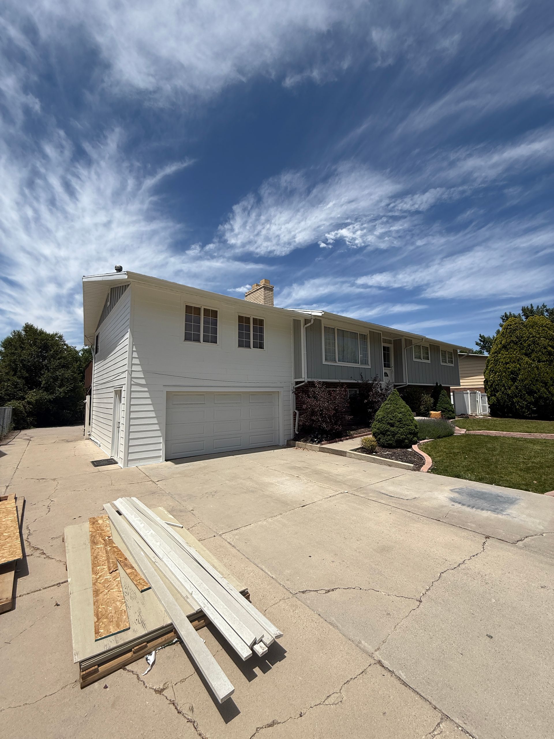 A white two-story house with a gray garage door and driveway under a blue sky with clouds.
