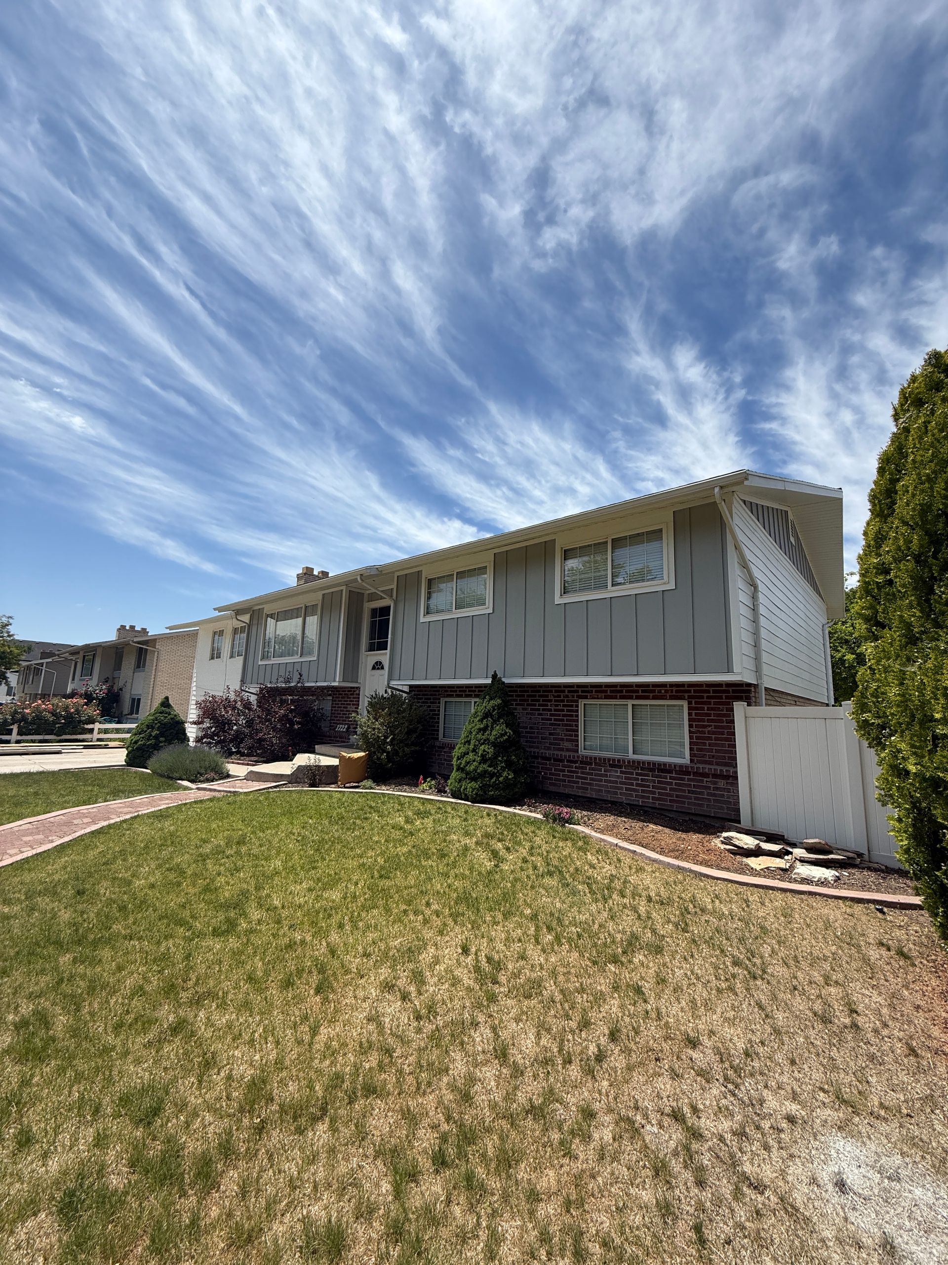 A two-story house with a gray upper level and a brick lower level under a blue sky with streaky clouds.