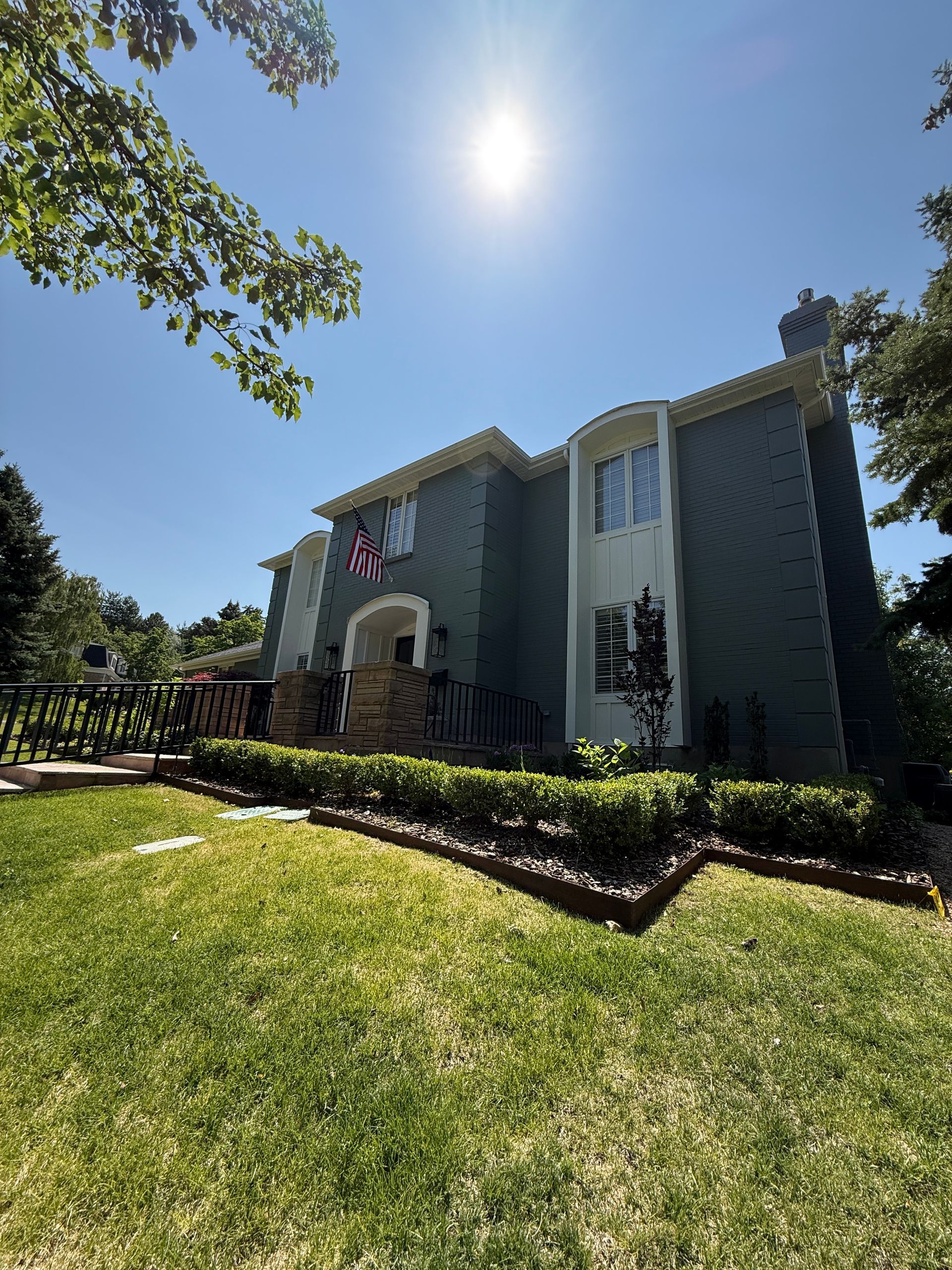 Two-story green house with manicured lawn, bright sun, and an American flag.