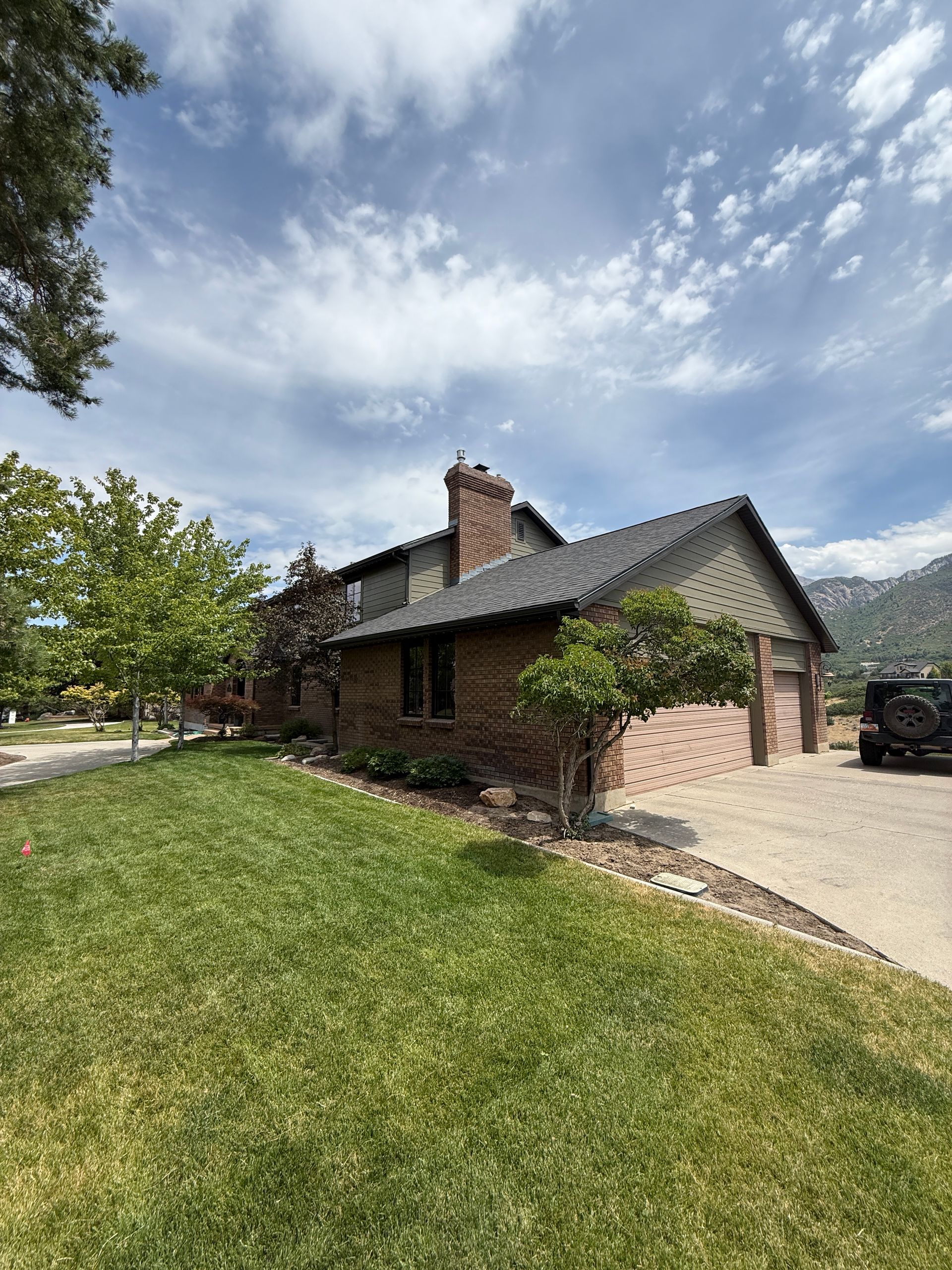 House with brick and siding, green lawn, driveway, cloudy sky, and trees.