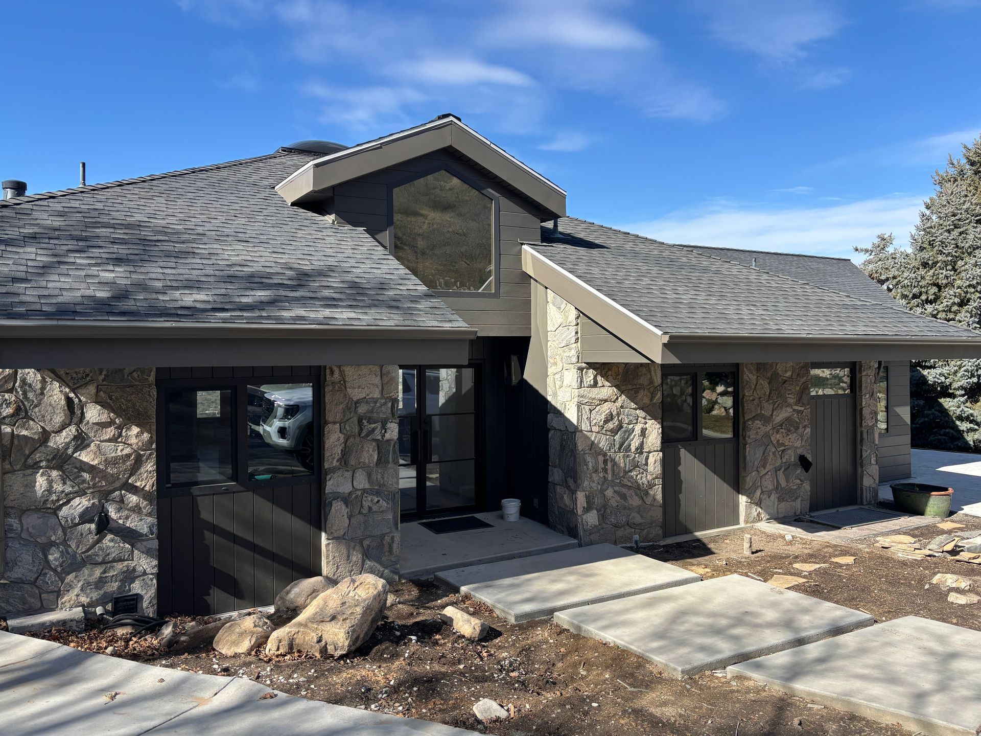 Stone-clad house with gray roof, black trim, and a concrete walkway leading to the front door on a sunny day.
