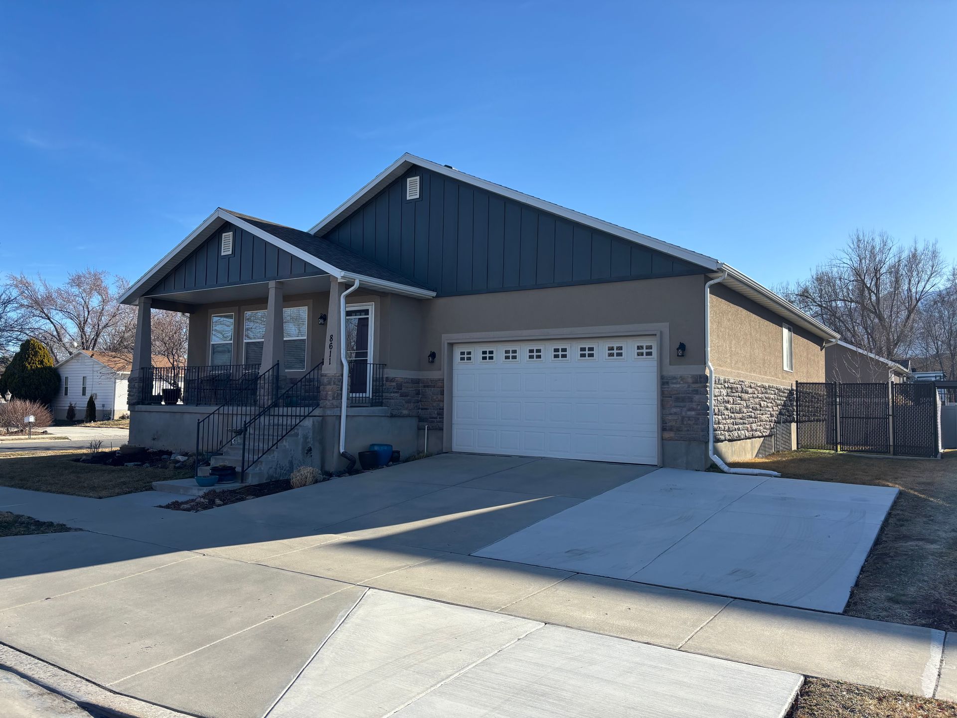 Single-story house with gray siding, stone accents, white garage door, and a concrete driveway under a clear blue sky.