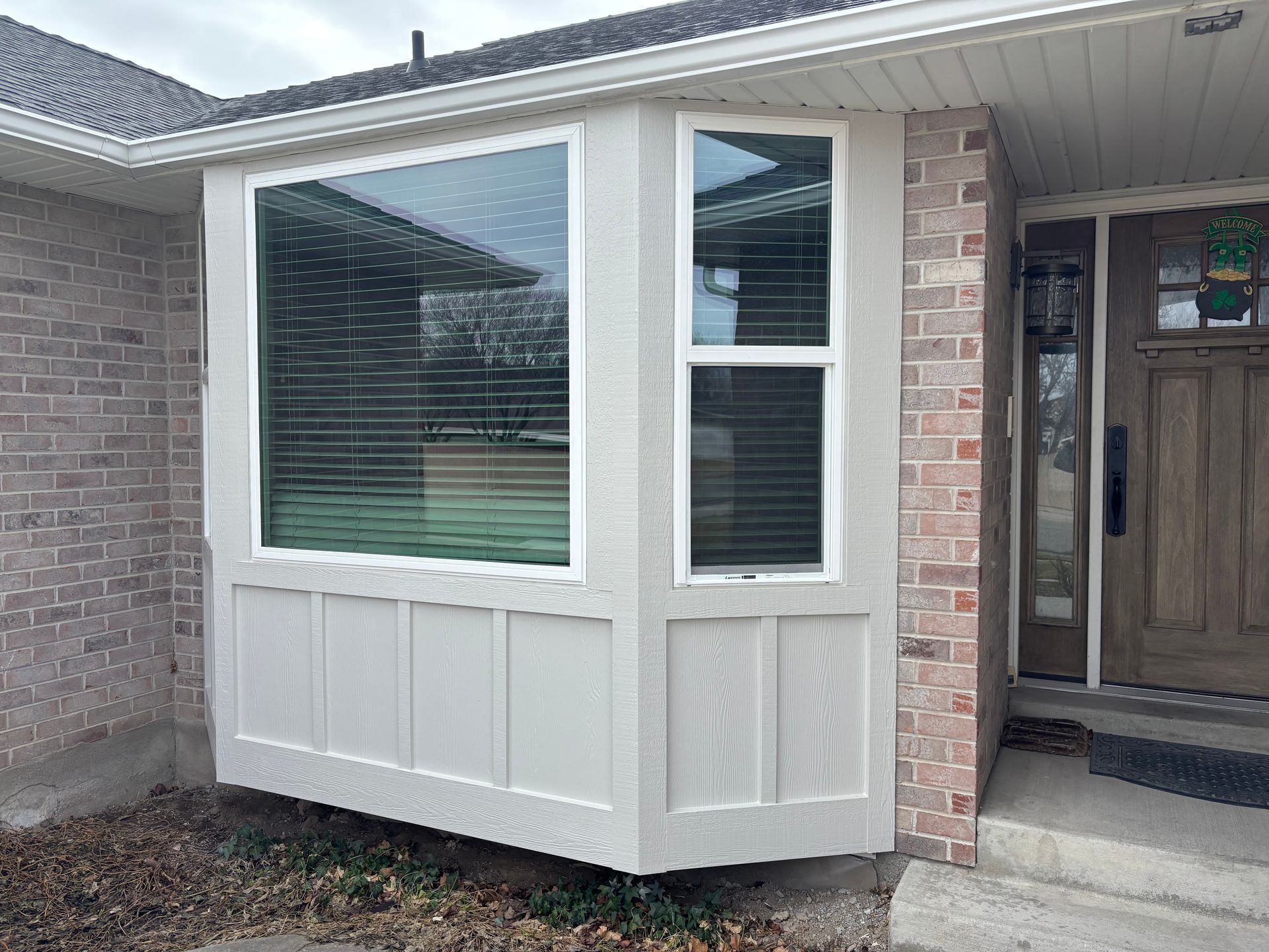 Bay window on a brick house with blinds, beige trim and a wooden door.