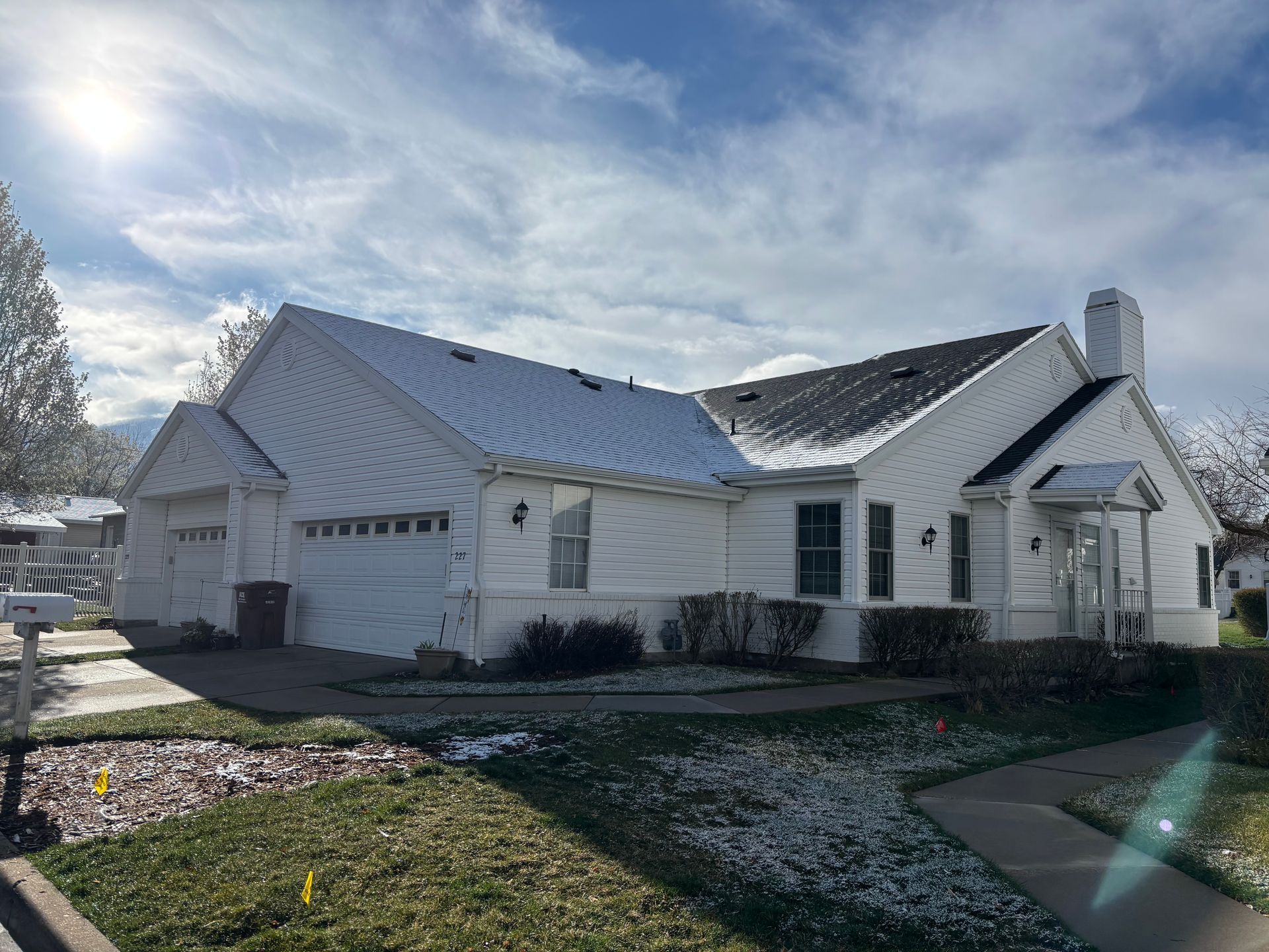 White house with two-car garage, snow-covered roof, and patches of grass in sunlight.