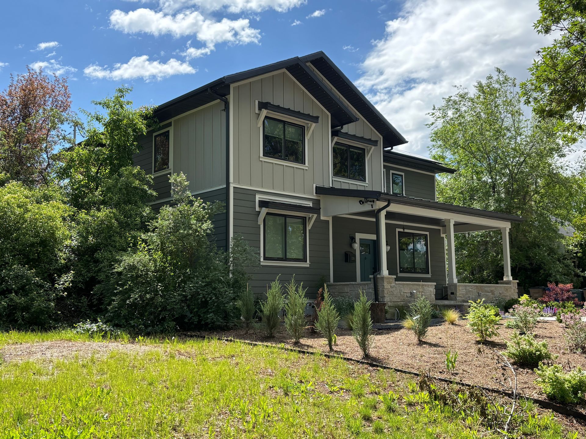 Two-story house with green siding, dark trim, and a covered porch, surrounded by greenery under a cloudy sky.