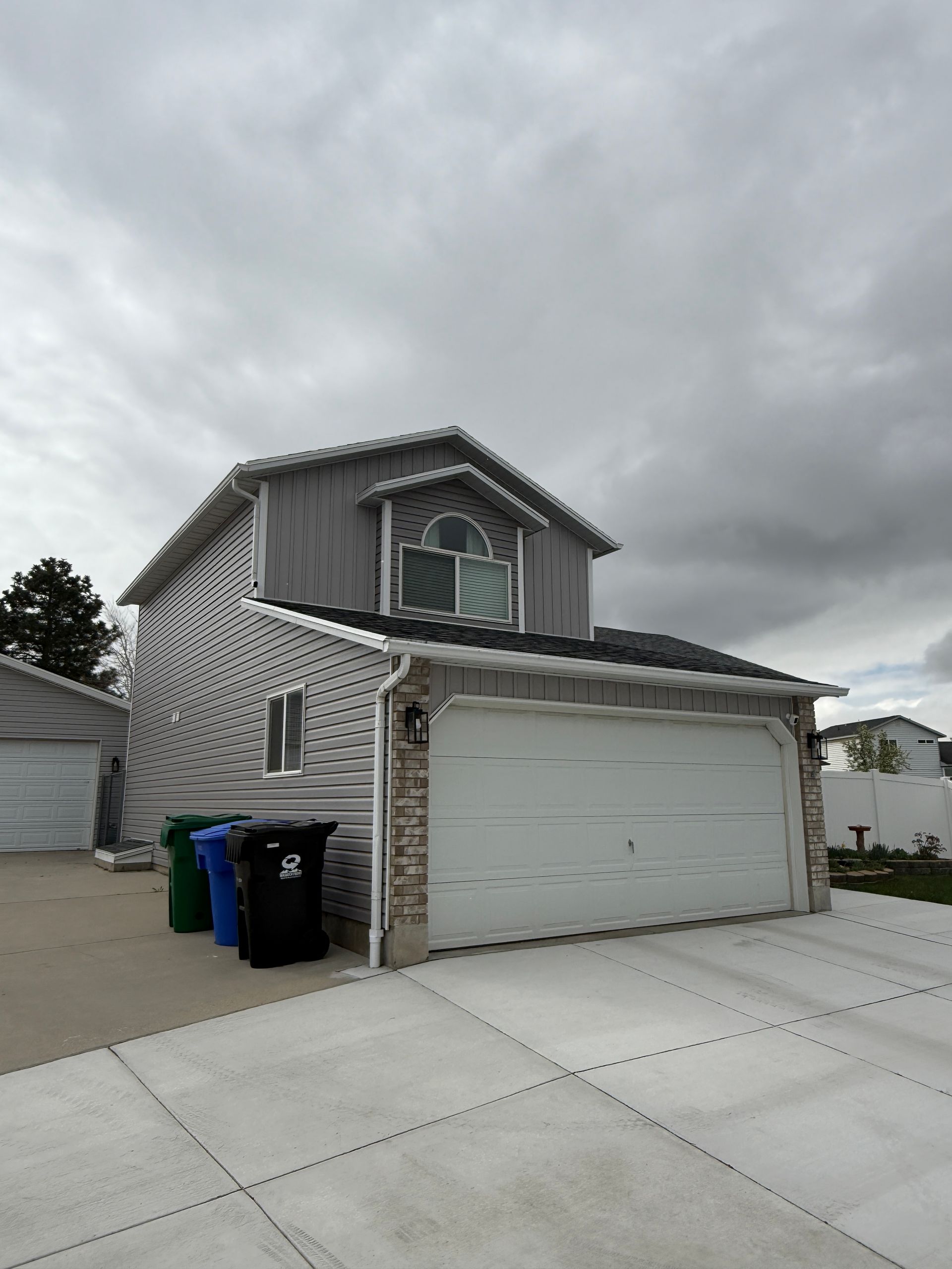 Two-story house with attached garage under a cloudy sky. Trash cans sit in the driveway.