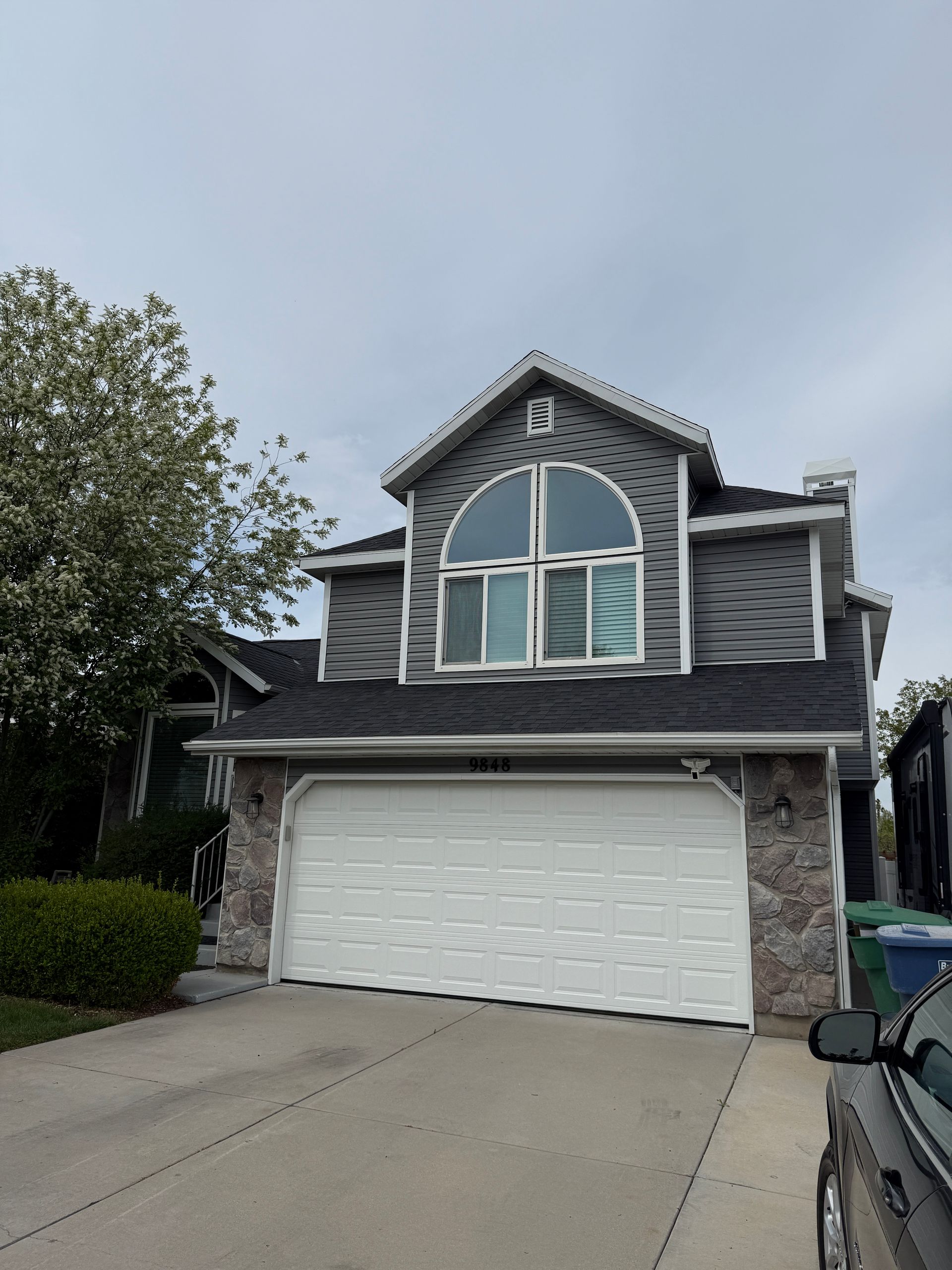 Two-story gray house with stone accents, white garage door, and large arched window under an overcast sky.