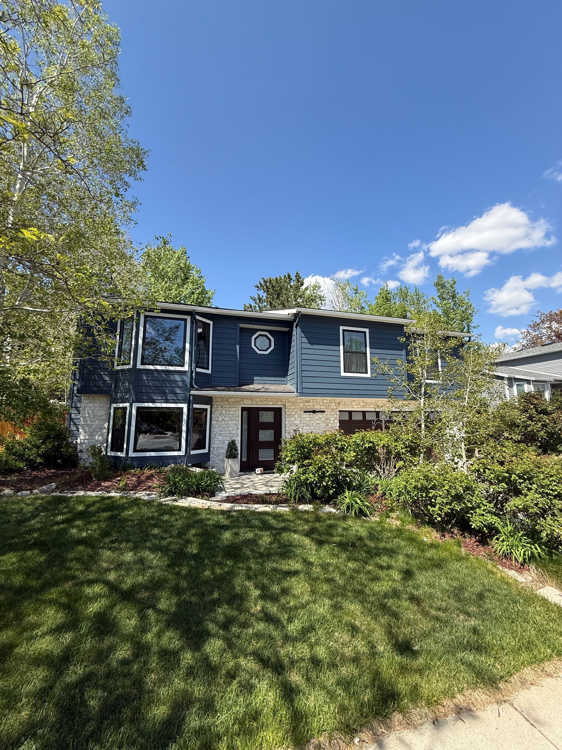 Two-story blue and white house with large windows, surrounded by trees and a green lawn on a sunny day.