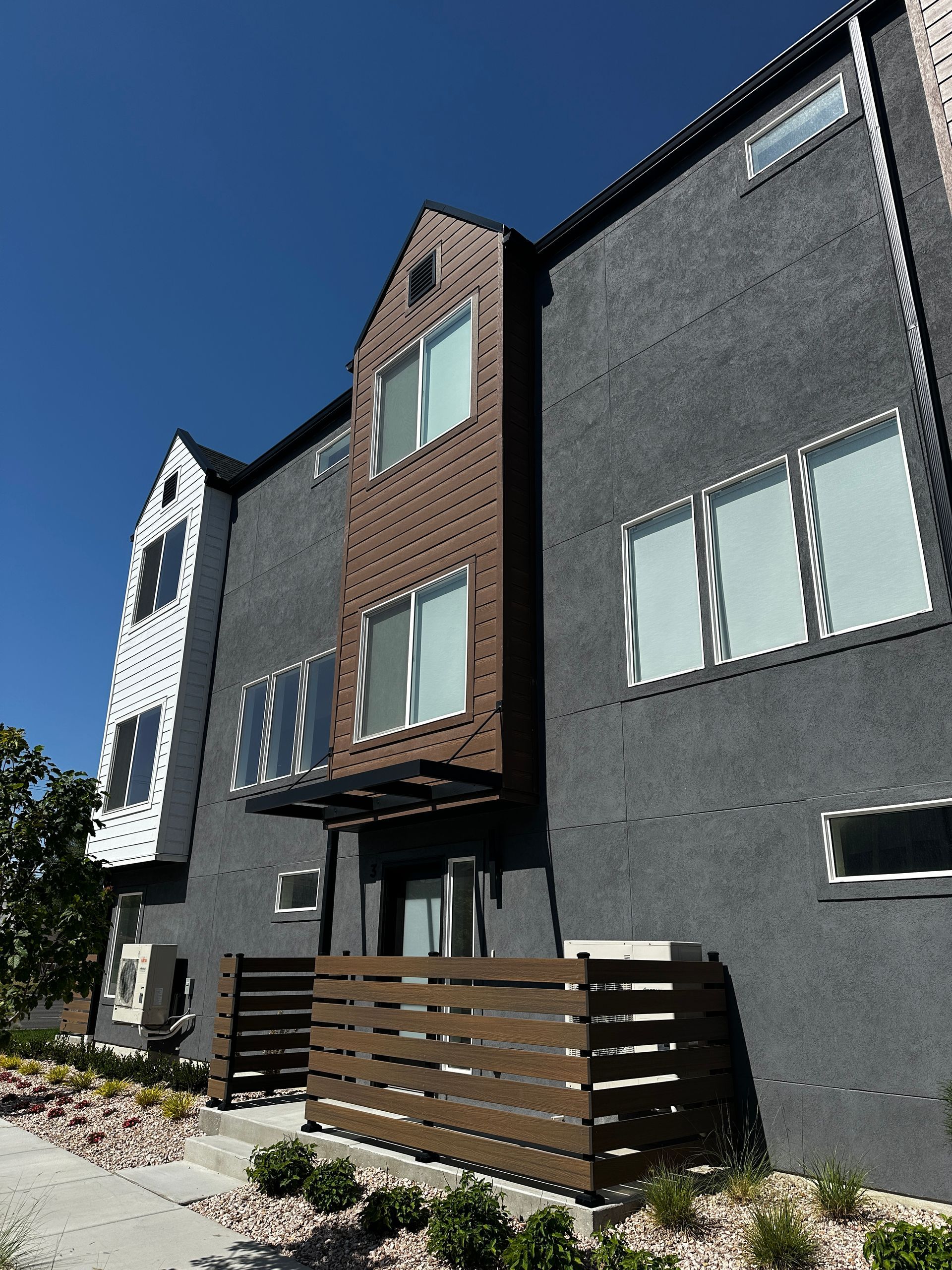 Three-story townhome with dark gray stucco and brown accent. Wooden fence and clear sky.
