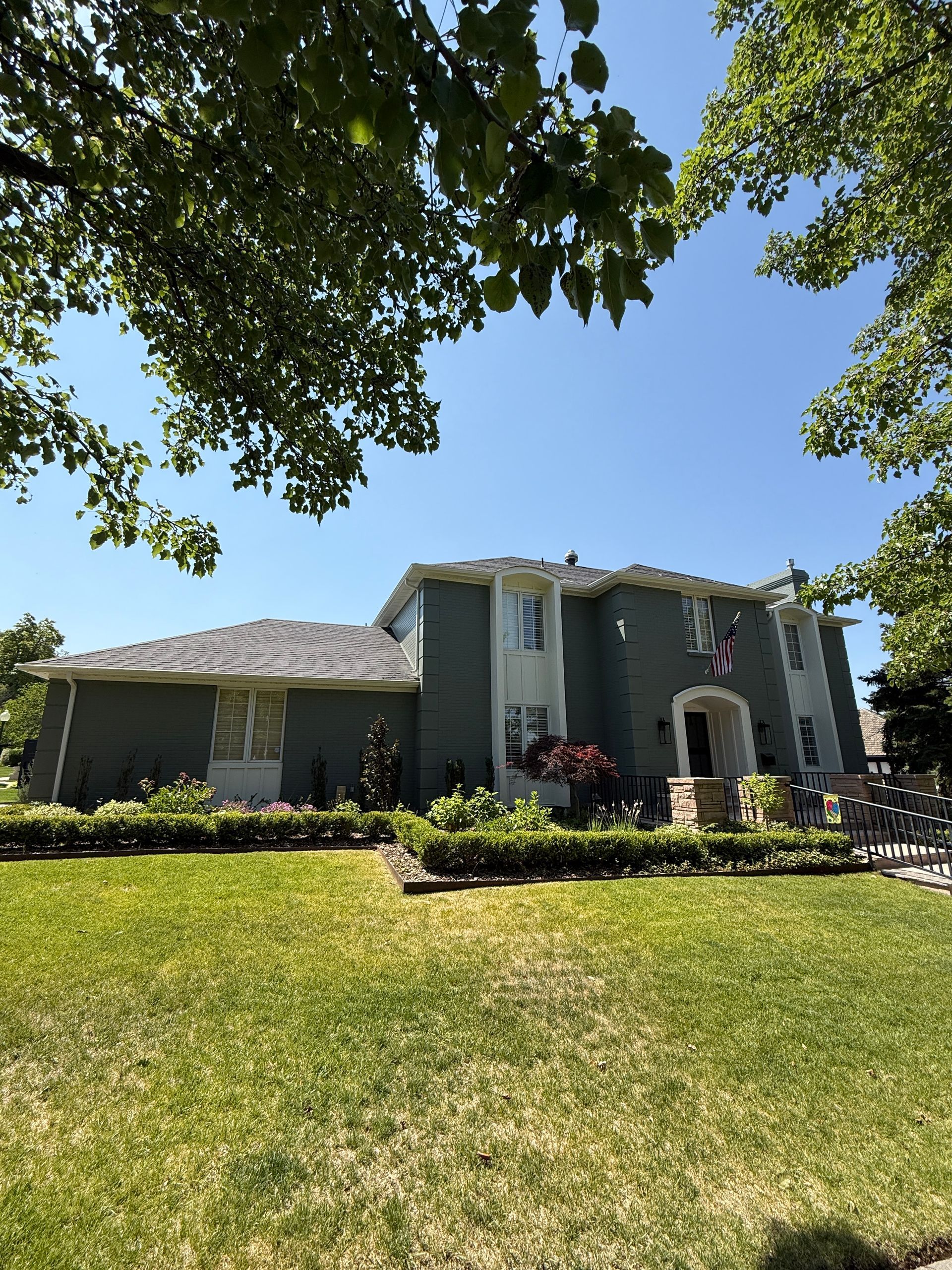 Two-story green house with white trim, green lawn, and trees under a blue sky.
