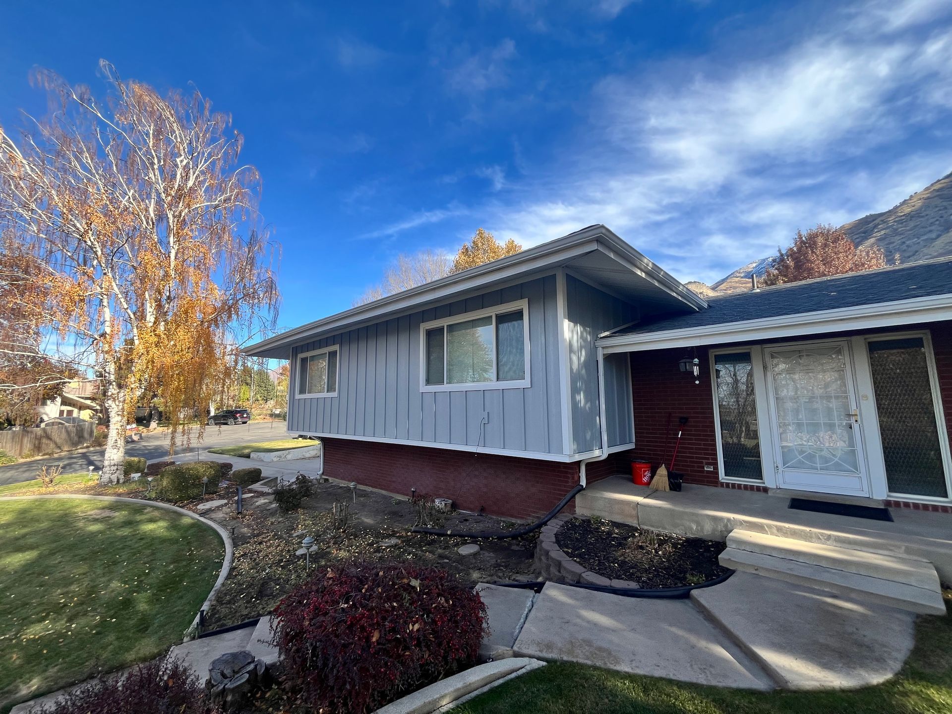 House with gray siding, red brick base, and autumn foliage under a blue sky.