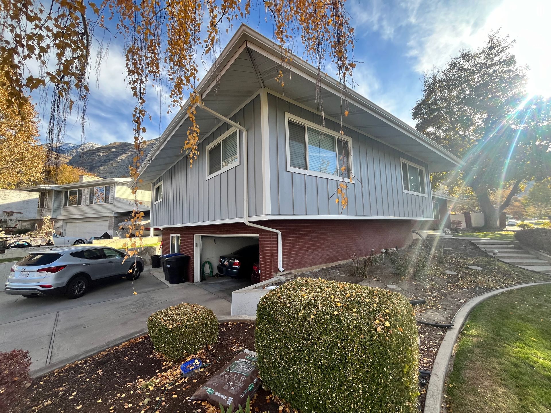 Two-story gray house with red brick base and attached garage. A car sits in the driveway on a sunny day.