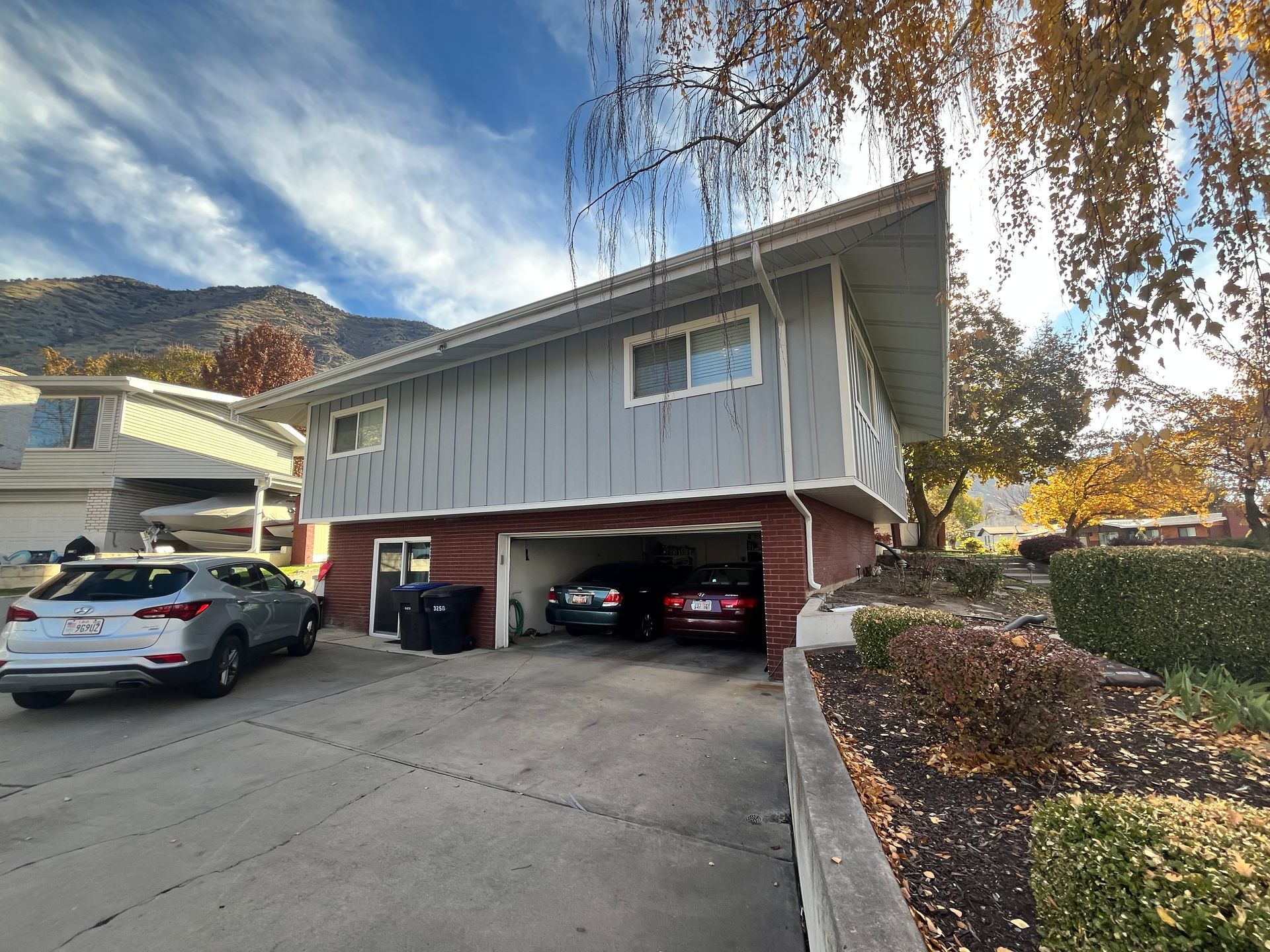 Two-story gray house with a garage, parked cars, and mountain backdrop.