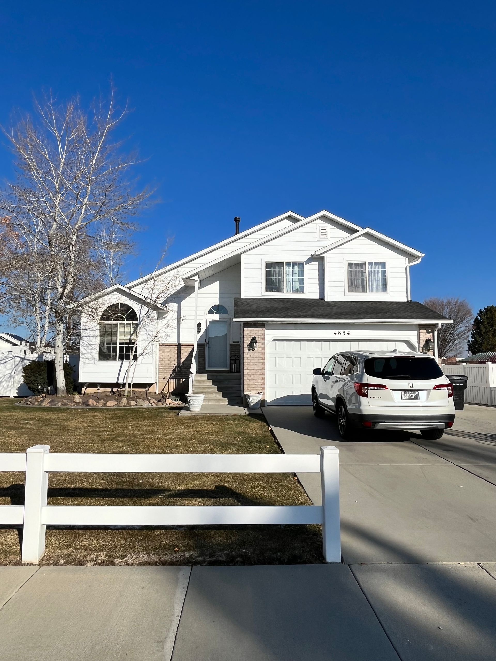 White two-story house with a car in the driveway, a white fence, and a clear blue sky.