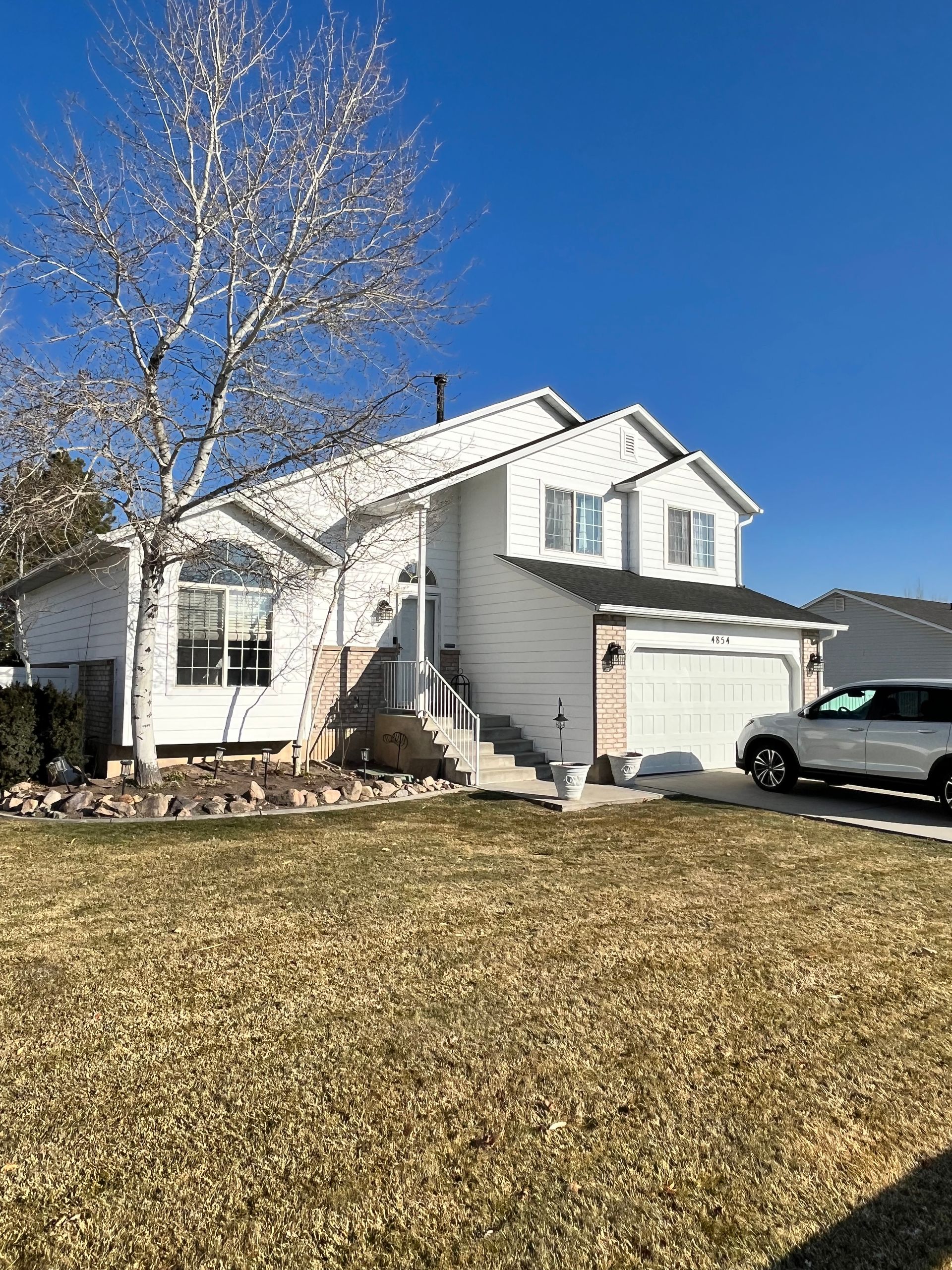 Two-story white house with garage, a bare tree, and a car parked in front, on a sunny day.