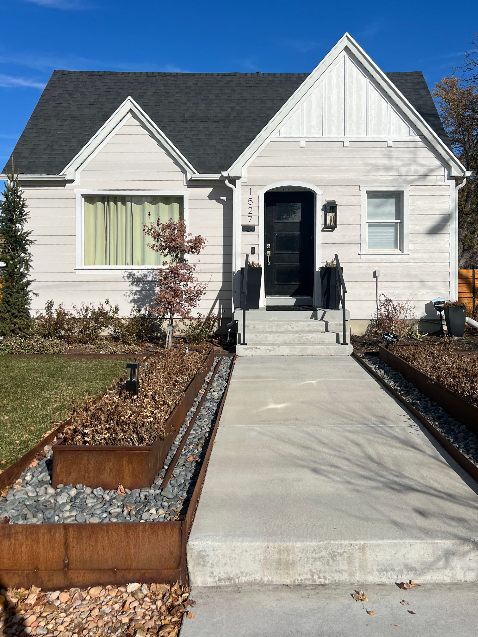 Tan house with black door, concrete path, and landscaped beds with dry leaves.