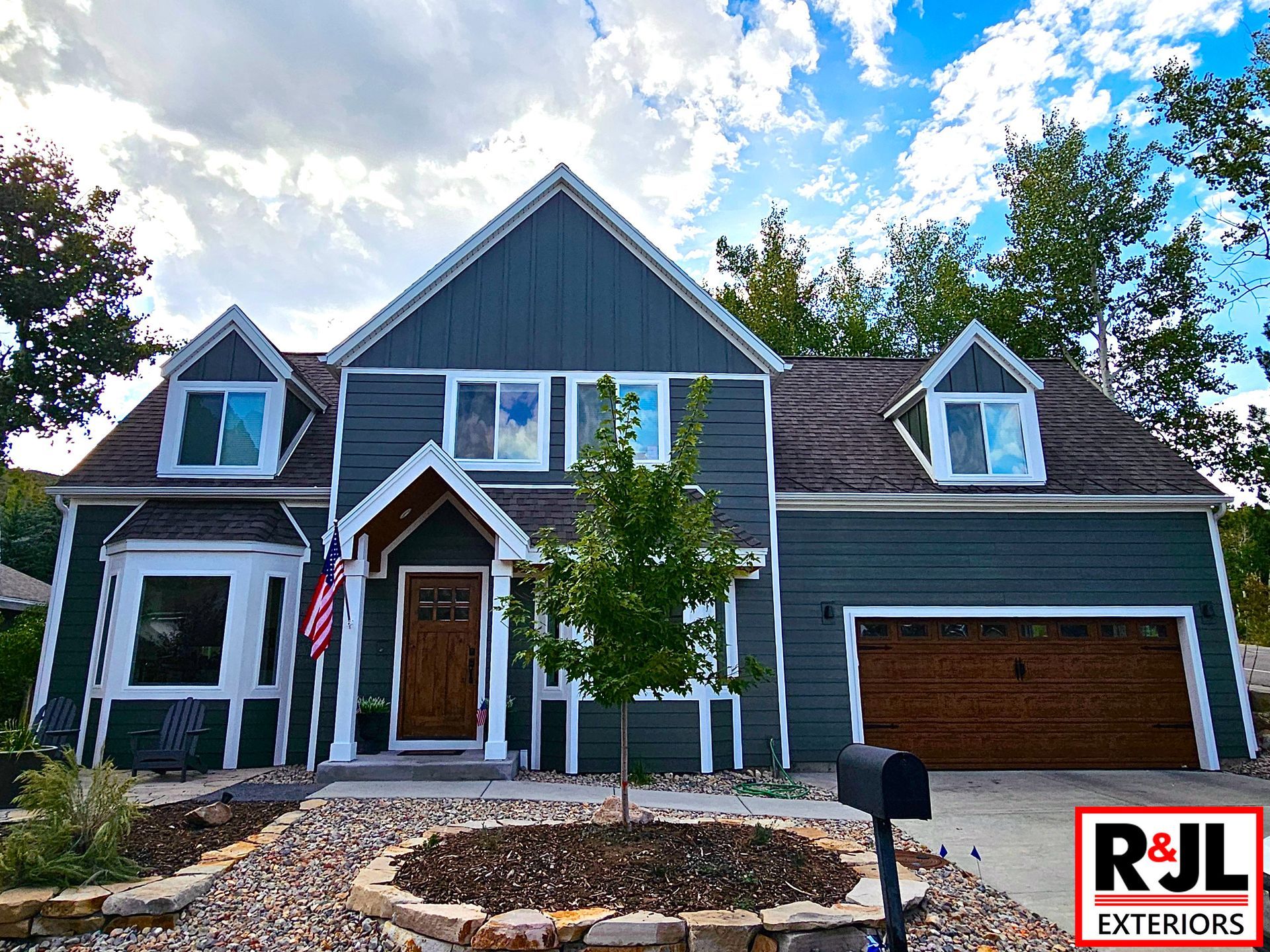 Two-story house with dark teal siding, brown garage door, and a circular flowerbed in the front yard.