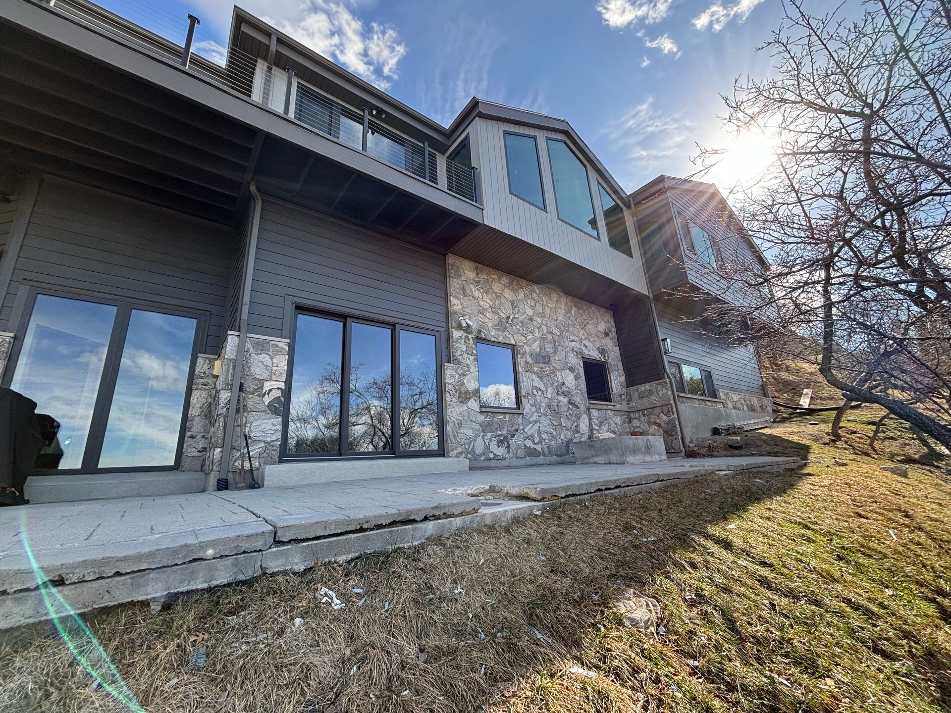 Two-story house with stone and gray siding, large windows, and a sunny, grassy slope.
