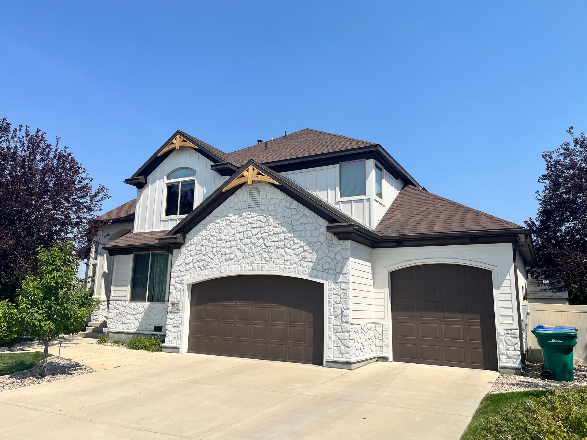 Two-story house with white textured exterior, brown roof, and garage doors. Green trash can on right.