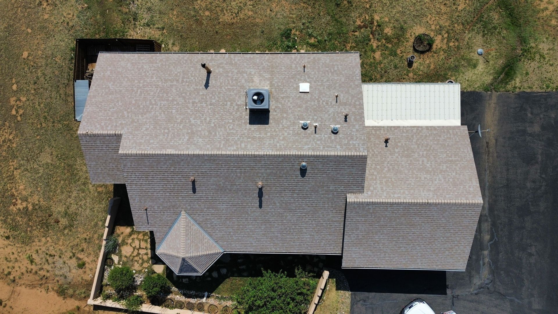 Overhead view of a house with a brown shingled roof, a black paved driveway, and surrounding dry vegetation.