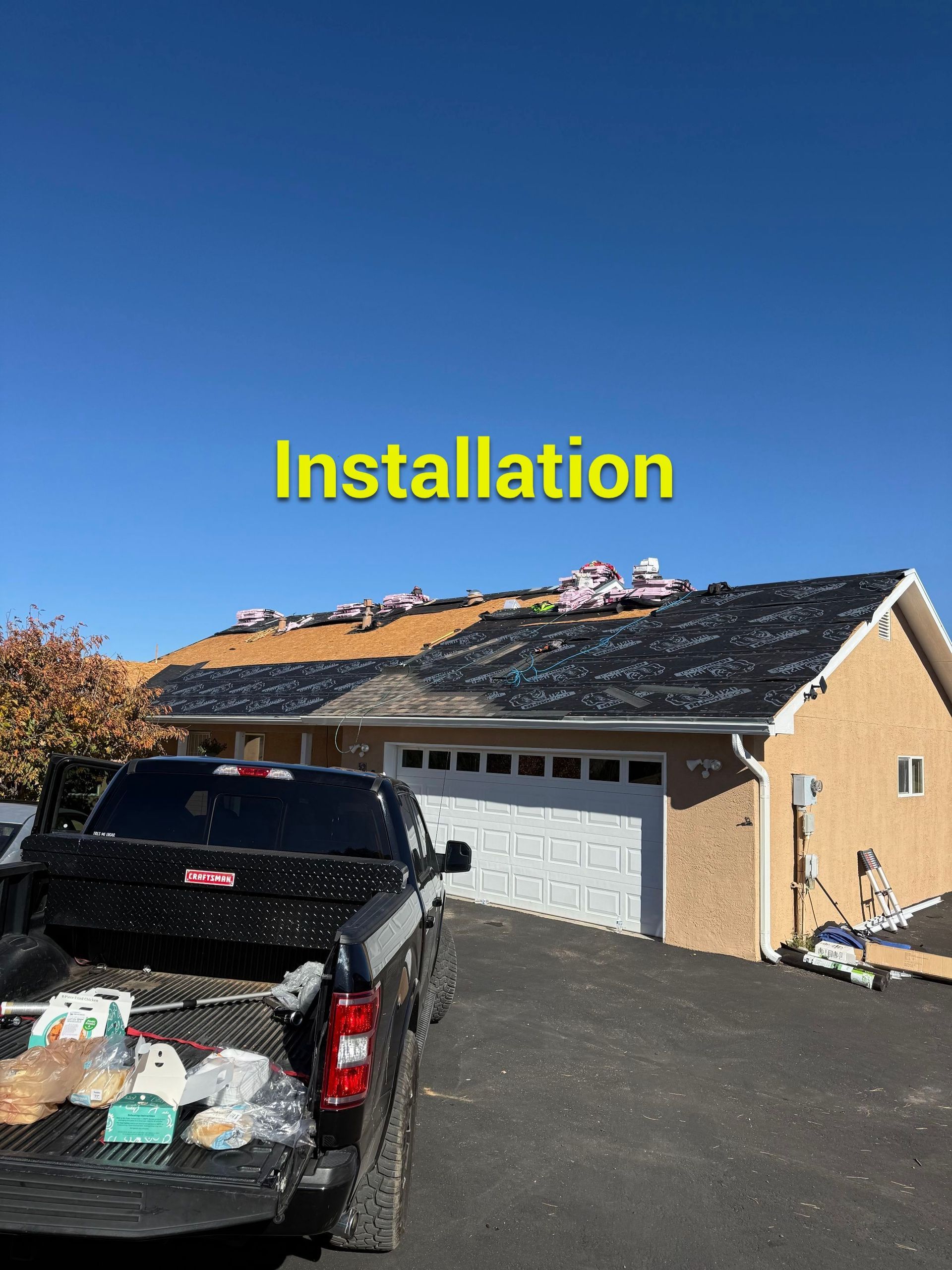 Black truck bed with roofing materials and house with roof installation underway.