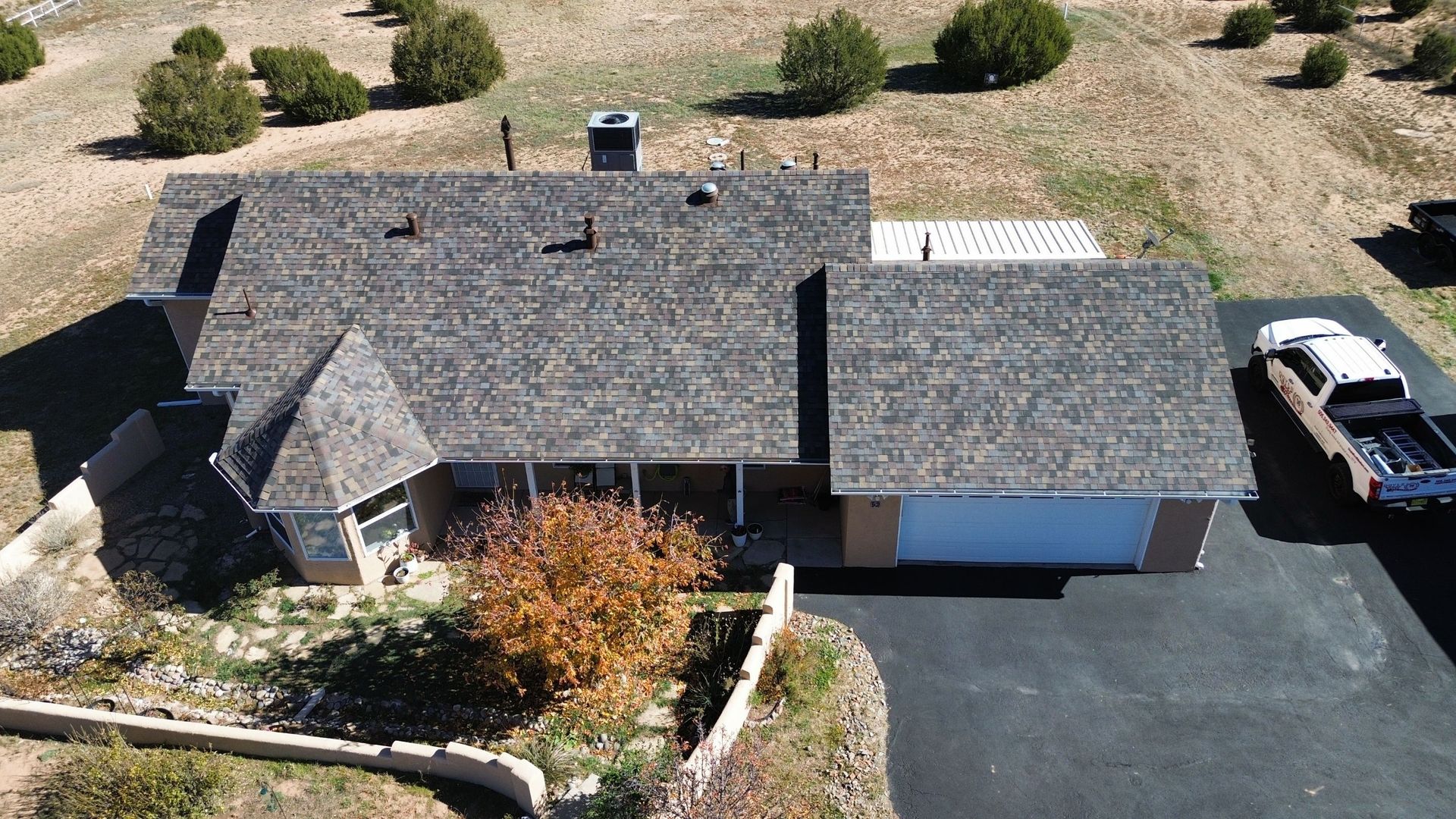 Aerial view of a single-story house with a driveway and a truck parked beside it, surrounded by dry land and trees.
