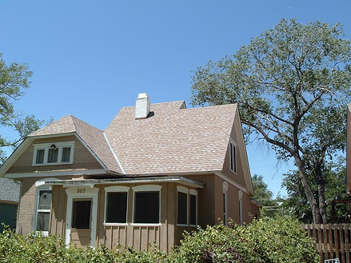 Tan house with gabled roof, small porch, and large tree in front. Blue sky background.