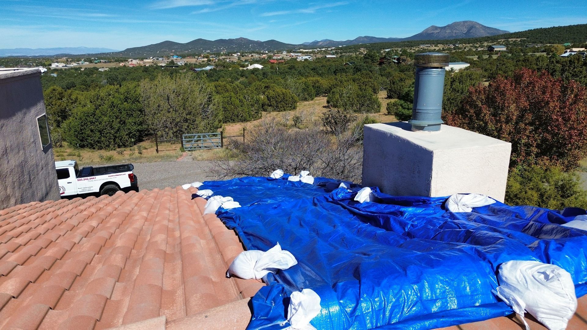 Roof covered with blue tarp, white bags, and a truck, overlooking a scenic landscape.