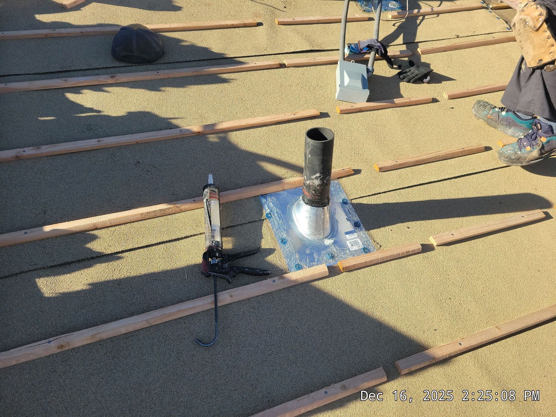 Roof with insulation, vent pipe, and tools. Sunlight creates shadows across wooden beams and granules.