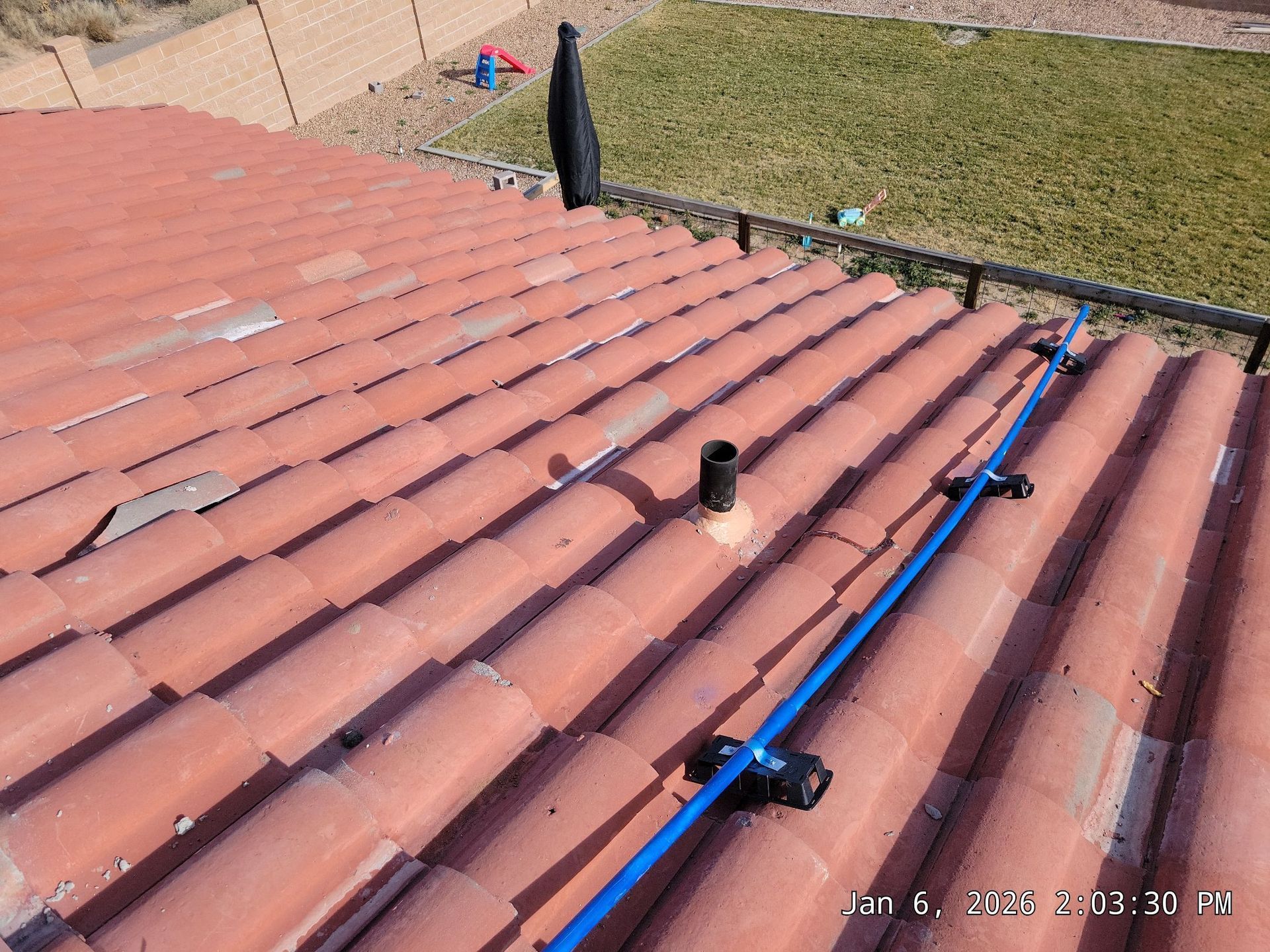 Red tile roof with a blue pipe secured by black brackets. Black vent pipe and a lawn are visible.