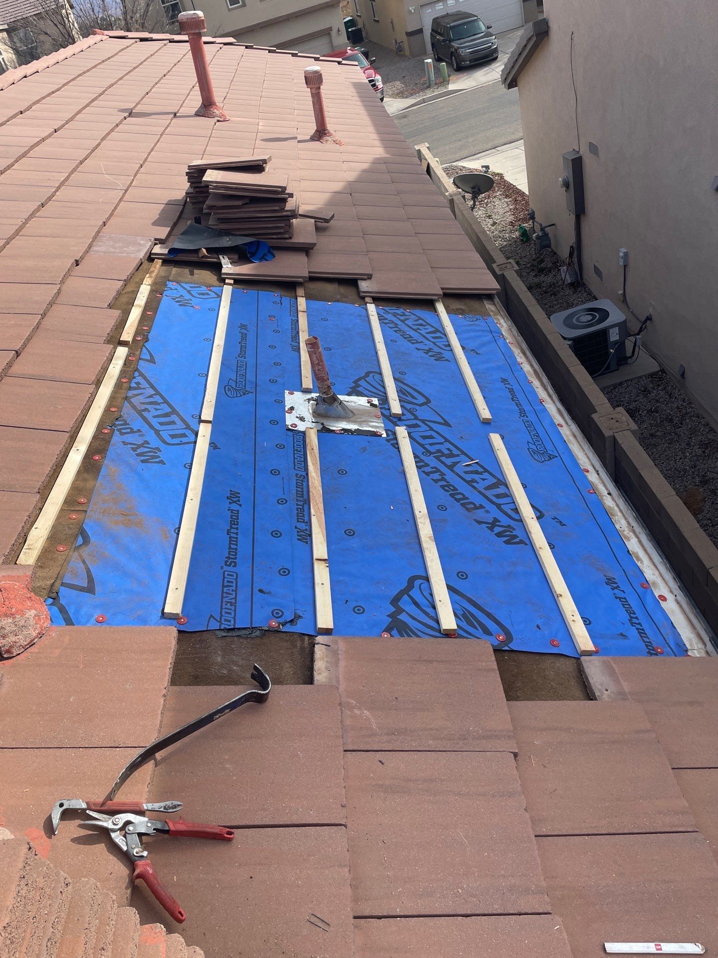 Roof partially dismantled, showing blue underlayment and wooden battens. Red tiles, tools, and vents visible.