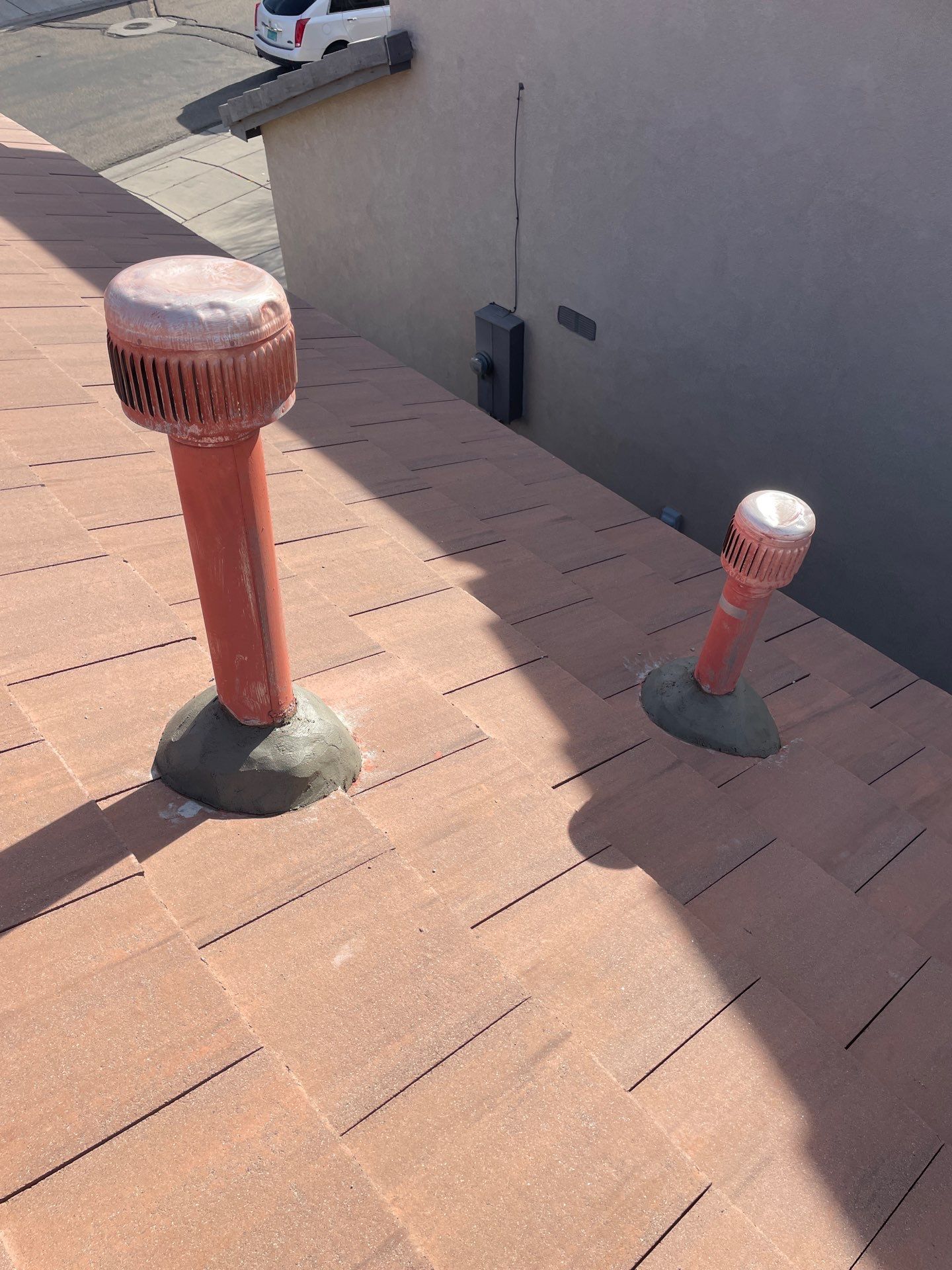 Two red roof vents on a brown shingled roof, next to a beige building with a small white vehicle in the background.