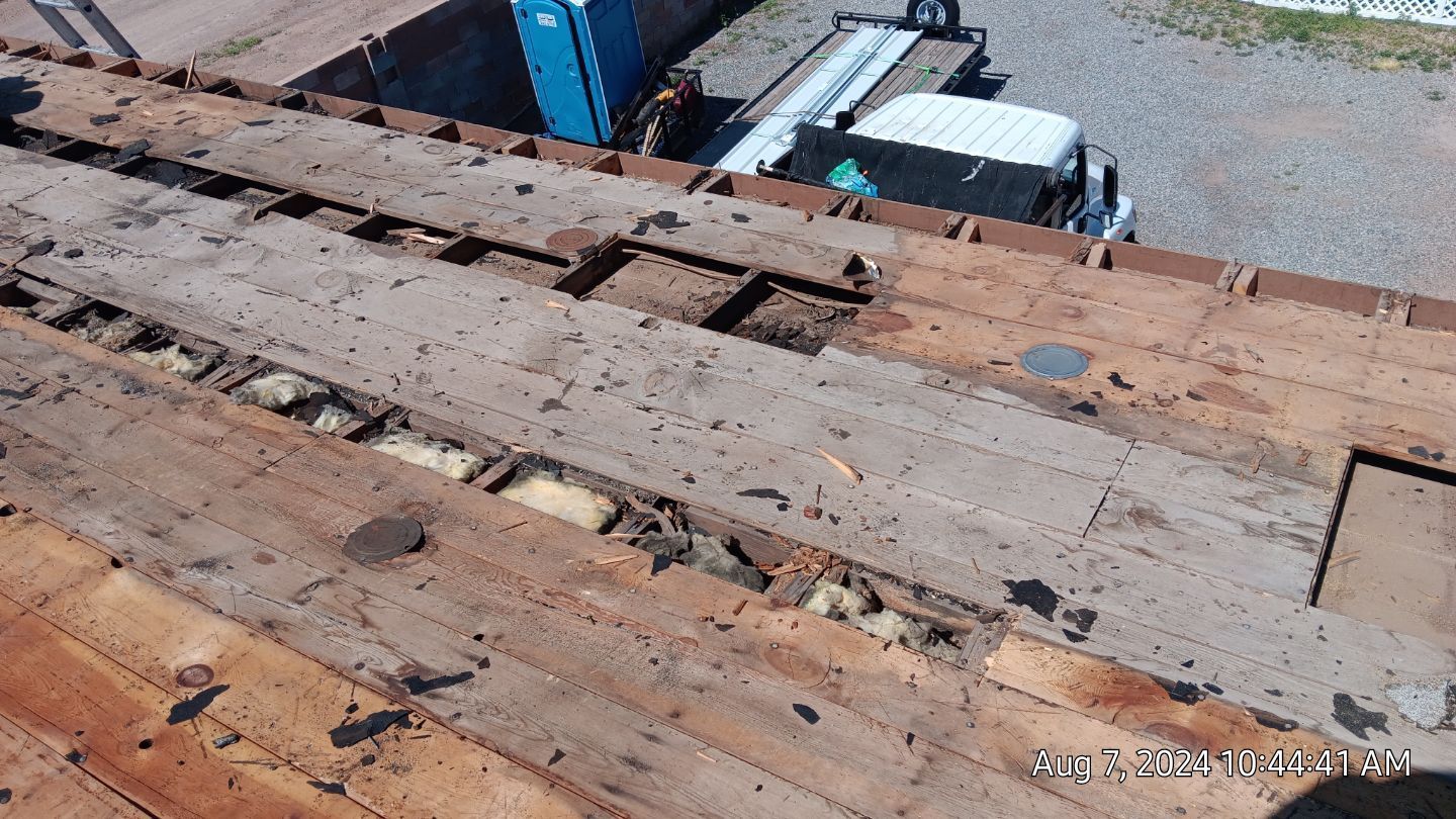 Damaged wooden roof with exposed insulation, construction equipment in background.