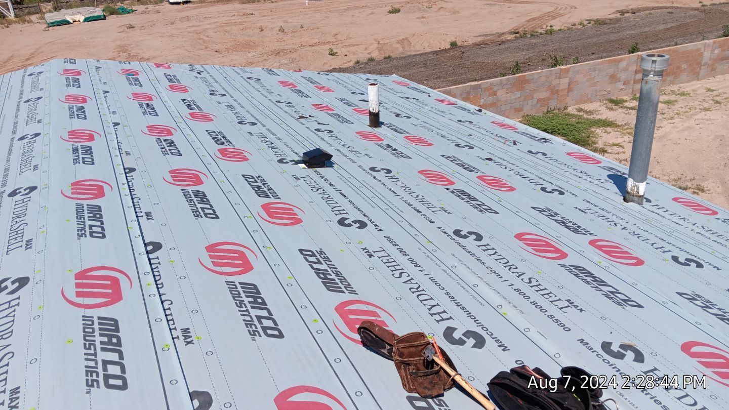 Rooftop with black underlayment, vent pipes, and tools in sunny outdoor setting.