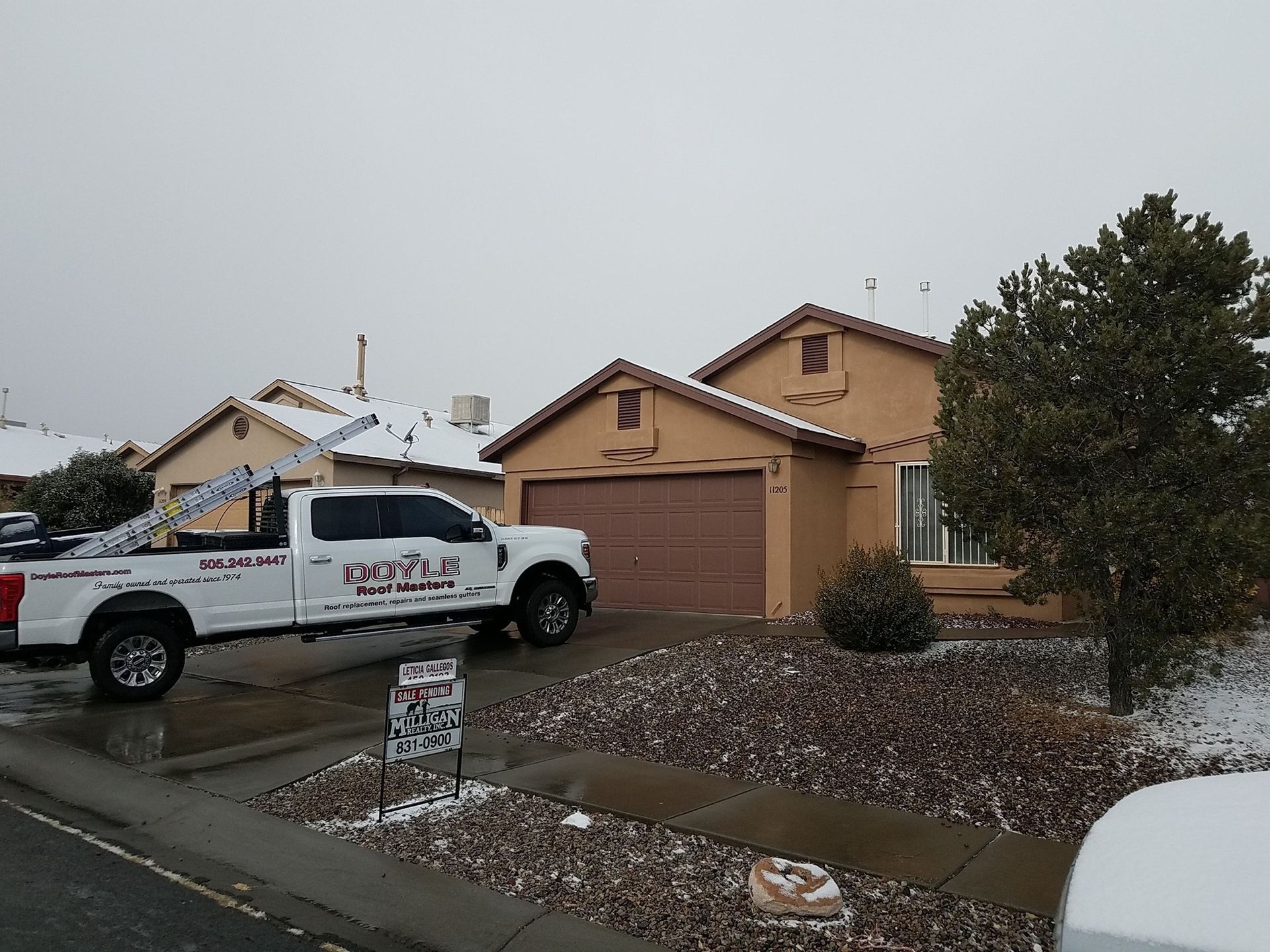 A white truck with a ladder parked in front of a tan house covered in snow.